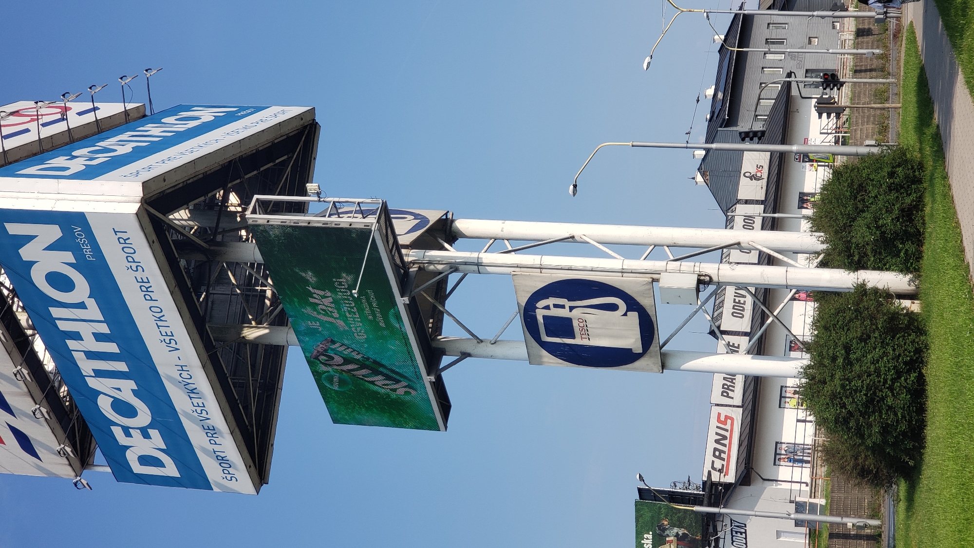 The image shows a street view in Prešov, Slovakia, on a sunny day. In the foreground, a sidewalk is visible with green grass and bushes. Dominating the upper part of the image is a large billboard for Decathlon, a sports retail company, with the word "PREŠOV" indicating the location. Below the Decathlon billboard, another digital billboard displays an advertisement for Sprite. Further down, a circular blue sign with a white gas pump symbol and the word "TESCO" is mounted on a white pole. In the background, a white building with black signage reads "CANIS PRAVE ODEVY", suggesting a clothing store. Adjacent to this building is a darker-colored building with solar panels on its roof. Traffic lights are also visible in the distance. A few people are present, including one person walking on the sidewalk in the lower right corner. The sky is clear and blue.