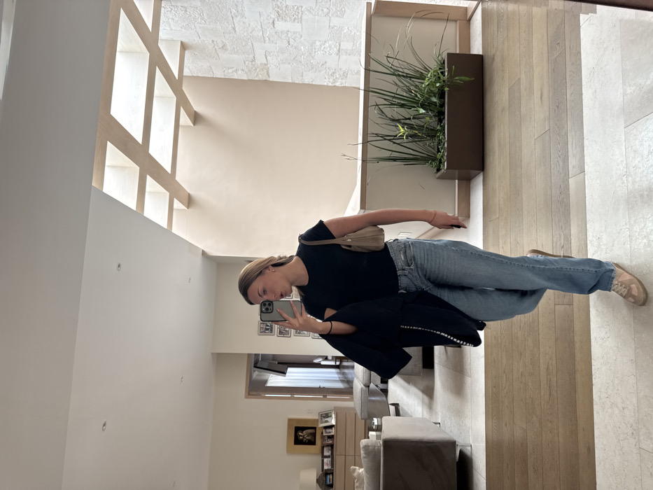A young woman takes a selfie in an upscale home. She wears a black t-shirt, light blue jeans, and sneakers. A blazer is draped over her arm. She holds her phone in her left hand, focusing on the screen. The interior features a high ceiling with grid detailing, light-colored walls, a wall planter with tall greenery, and tiled floors. The living room is visible in the background, furnished with sofas and a bookshelf.
