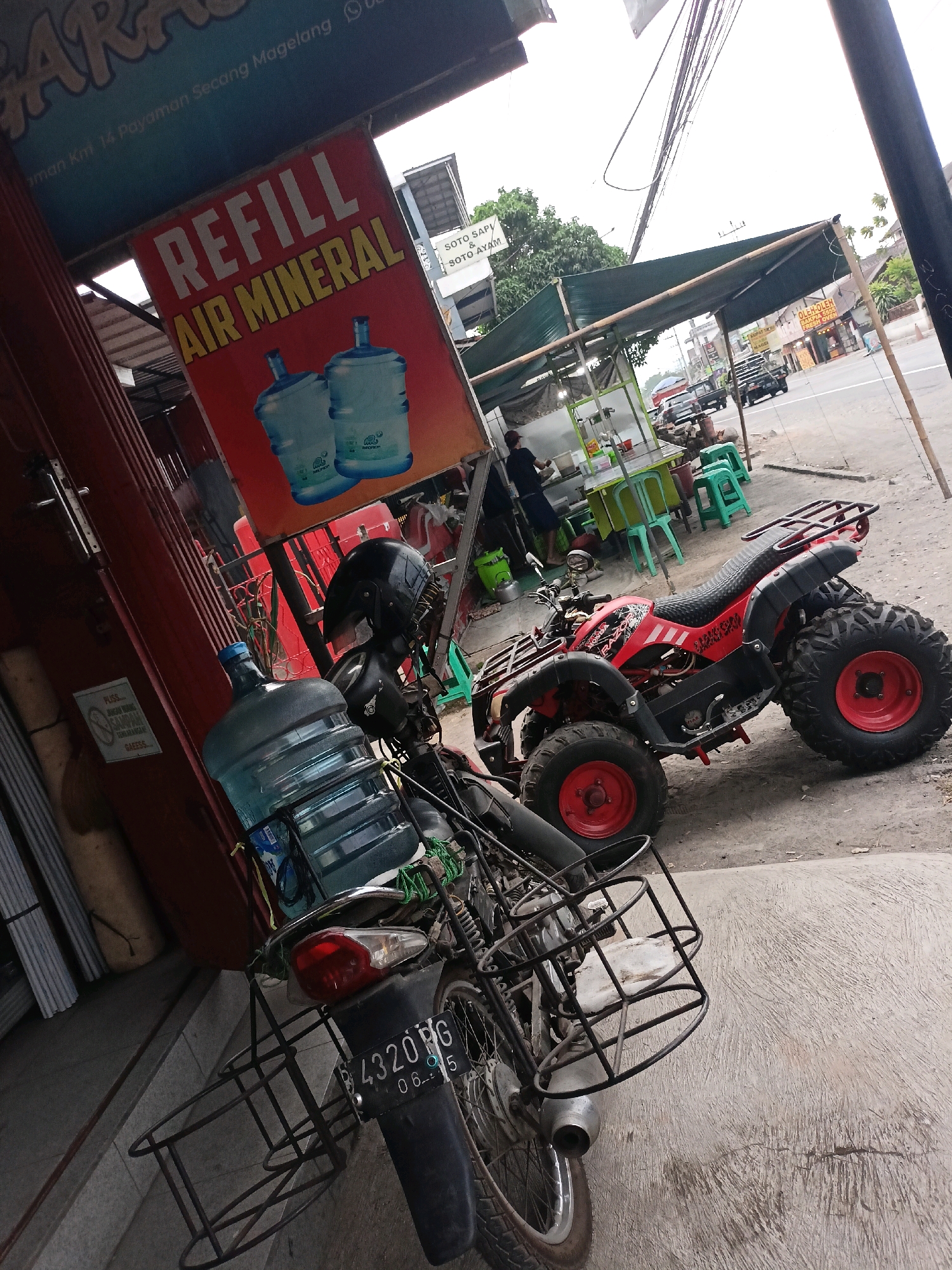 A motorbike with a large water jug strapped to its rear is parked on a sidewalk in Payaman, Indonesia, next to a red and black all-terrain vehicle (ATV). A sign above the motorbike advertises "REFILL AIR MINERAL". In the background, a street vendor stall with green tables and chairs is set up under a green awning, with several vehicles passing on the road. The scene appears to be a roadside business area.