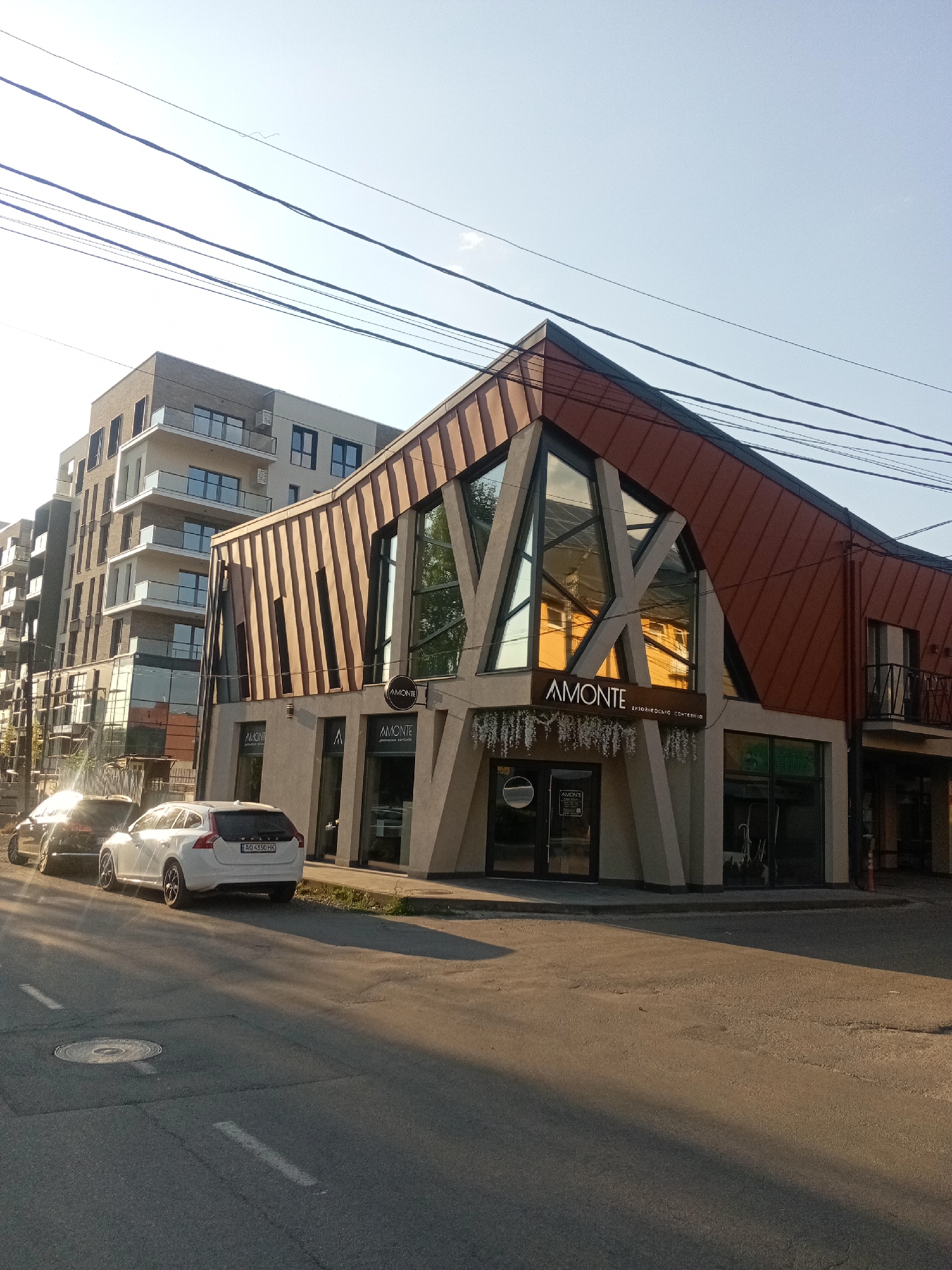 The image shows a street scene in Tiachiv, Ukraine, featuring a modern, angular building with large glass windows and a copper-colored roof. The building has the name "AMONTE" prominently displayed. Adjacent to it is a taller, multi-story residential building with balconies. Two cars are parked on the street in front of the modern building. The sun is low in the sky, casting long shadows and creating a warm, golden light. Overhead, electrical wires are visible against a clear sky. The street surface is asphalt with painted lane markings and a manhole cover.