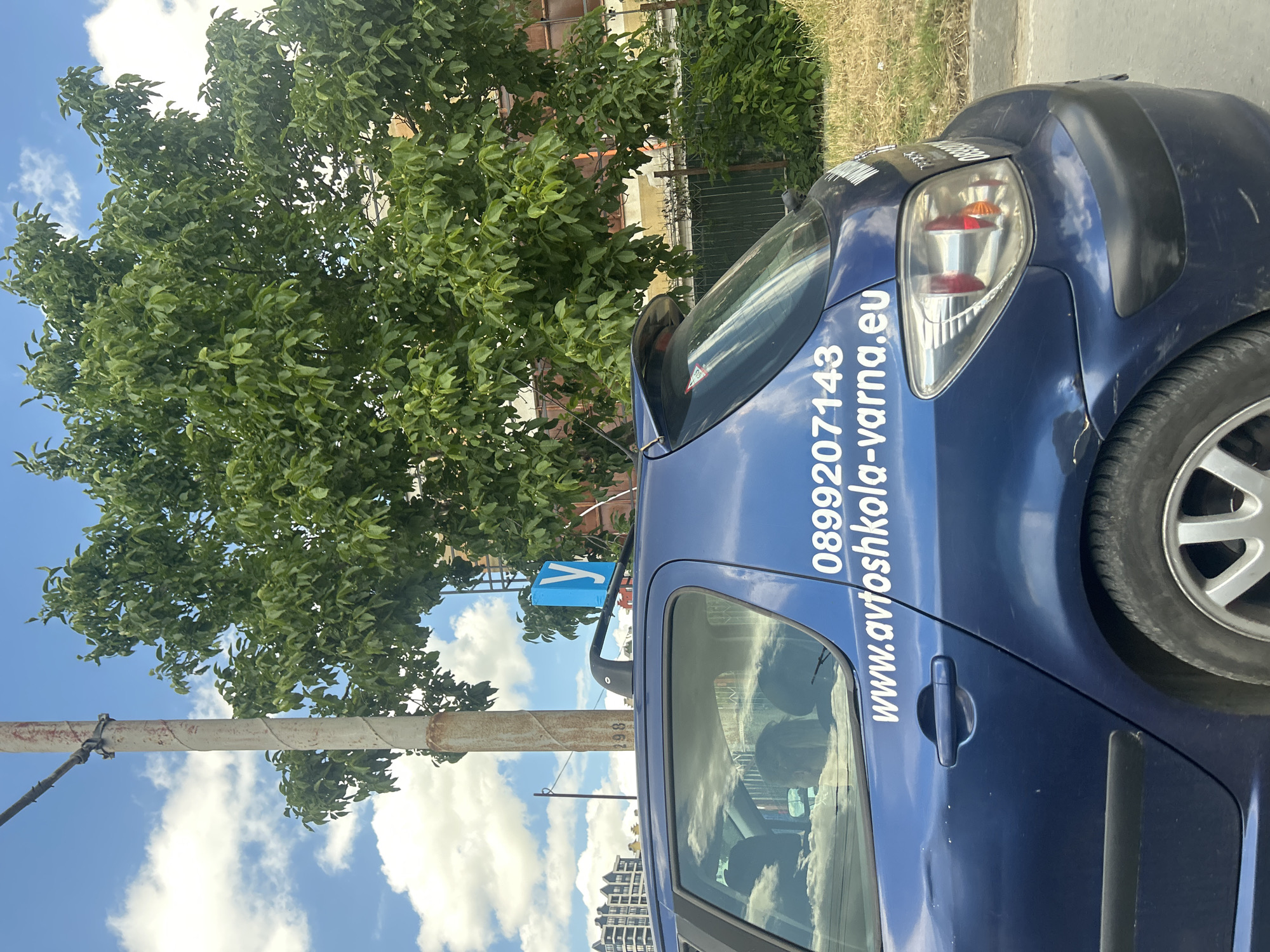 The image depicts a blue Peugeot 307 hatchback parked on a street in Varna, Bulgaria. The car has a roof rack with a bright blue sign that reads "У", a Cyrillic letter often used in Bulgaria to denote "Y" or "U", and commonly associated with driving schools. White lettering on the car's rear door displays a phone number, "0899207143", and a website address, "www.avtoshkola-varna.eu". In the background, a large, leafy tree dominates the scene, with buildings and a cloudy sky visible above. A utility pole with some wires is also present to the left of the car. The car appears to be in a residential or commercial area, with a fence and dry grass visible in the immediate background. The lighting suggests it is daytime.