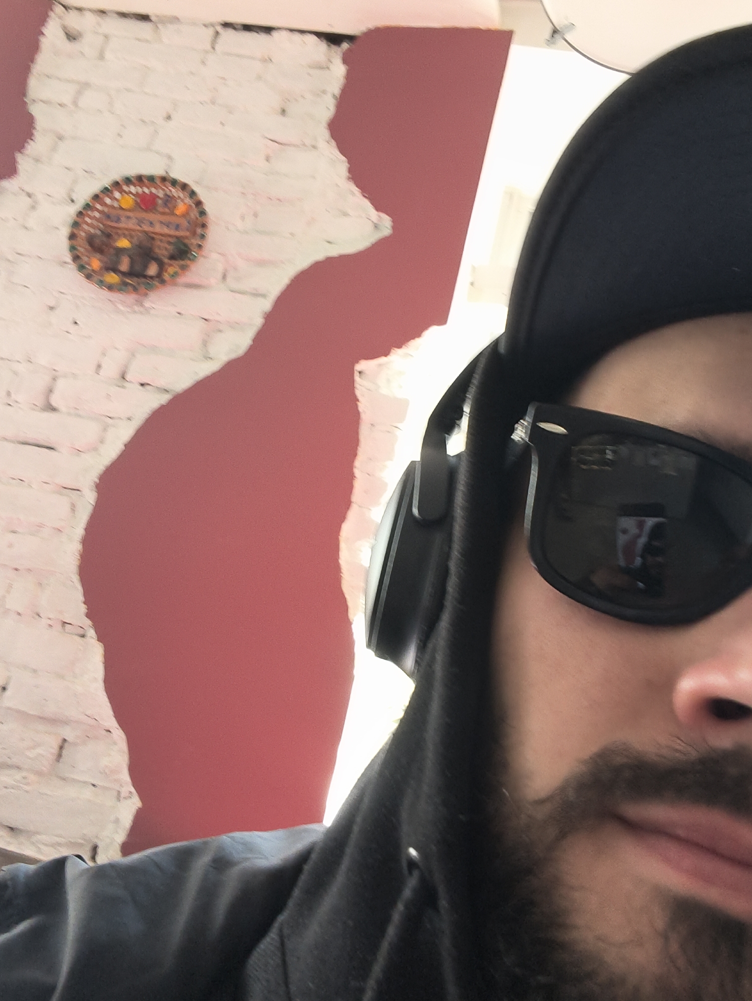 A selfie photograph shows a person wearing a black baseball cap, black sunglasses, and headphones. The person has a beard and mustache. The background features a brick wall partially painted pink and an ornamental circular decoration affixed to the wall. The location is Hrodna, Belarus.