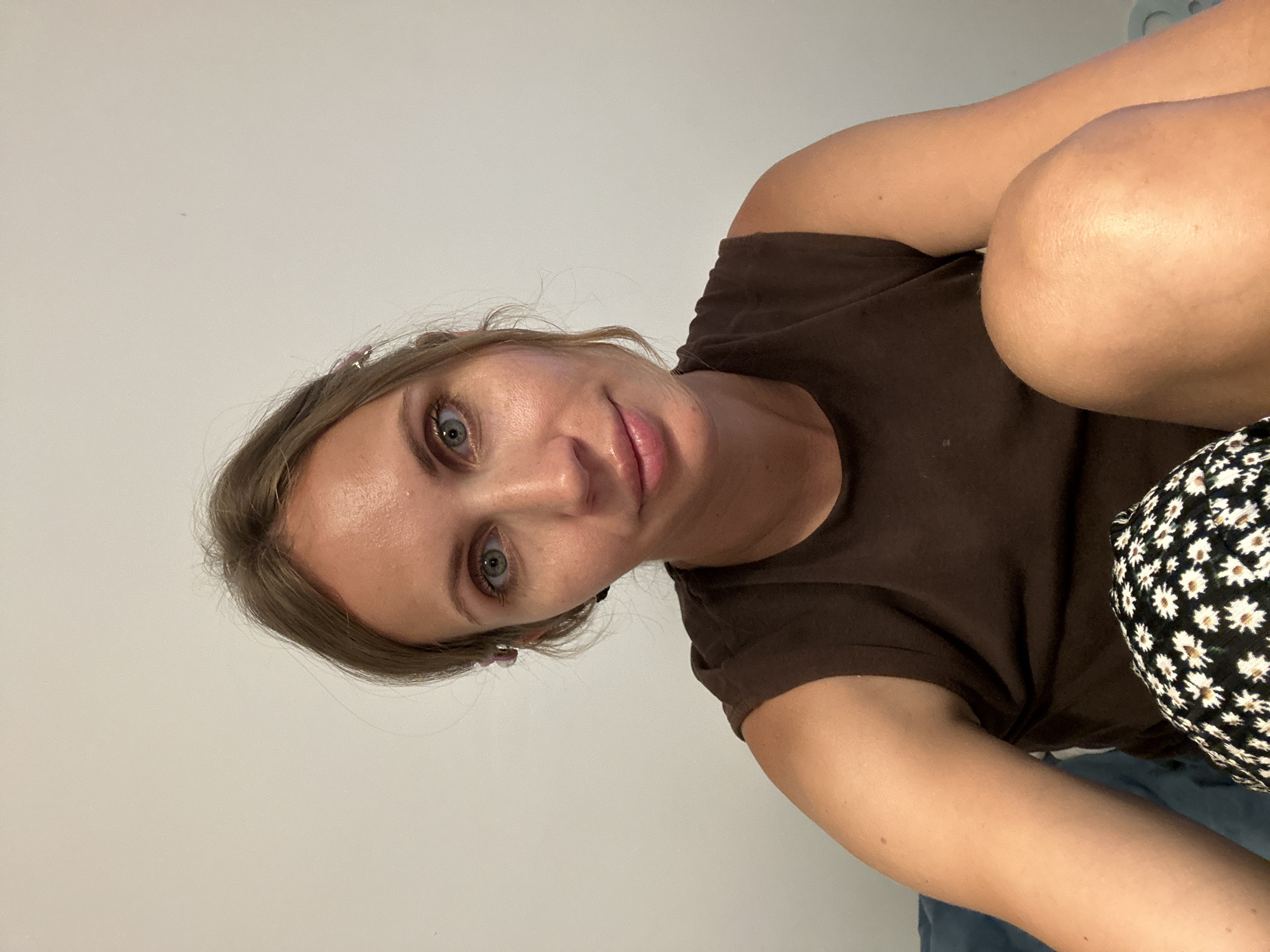 A woman with light brown hair and blue-green eyes is looking directly at the camera. She has fair skin and is wearing a brown t-shirt. Part of her right knee and leg is visible on the right side of the frame, and she appears to be wearing black and white floral patterned shorts or pants. The background is a plain white wall. There is no indication of the location being Fethiye, Turkey.