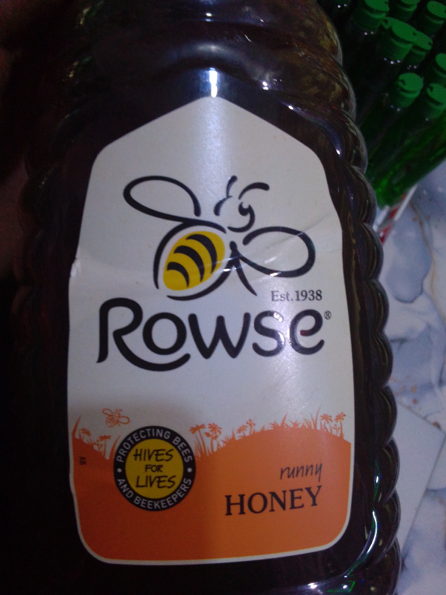 The image shows a close-up of a dark brown bottle of Rowse Runny Honey. The label is white with a black bee illustration at the top. Below the bee, the brand name "Rowse" is prominently displayed in large black lettering, with "Est. 1938" above it. The bottom portion of the label is orange and features the words "runny HONEY". There is also a circular emblem on the left side of the orange section that reads "PROTECTING BEES FOR LIVES AND BEEKEEPERS" with "HIVES FOR LIVES" in the center. The bottle appears to be sitting on a surface that is partially visible and is textured with what looks like fabric. In the background, to the right of the bottle, several green bottles with green caps are visible, suggesting they may be part of a larger product display or packaging. The lighting in the image is somewhat dim, with reflections visible on the glossy surface of the honey bottle. The image does not contain any information or visual cues that would indicate its location in the city of Howayi, Nigeria.