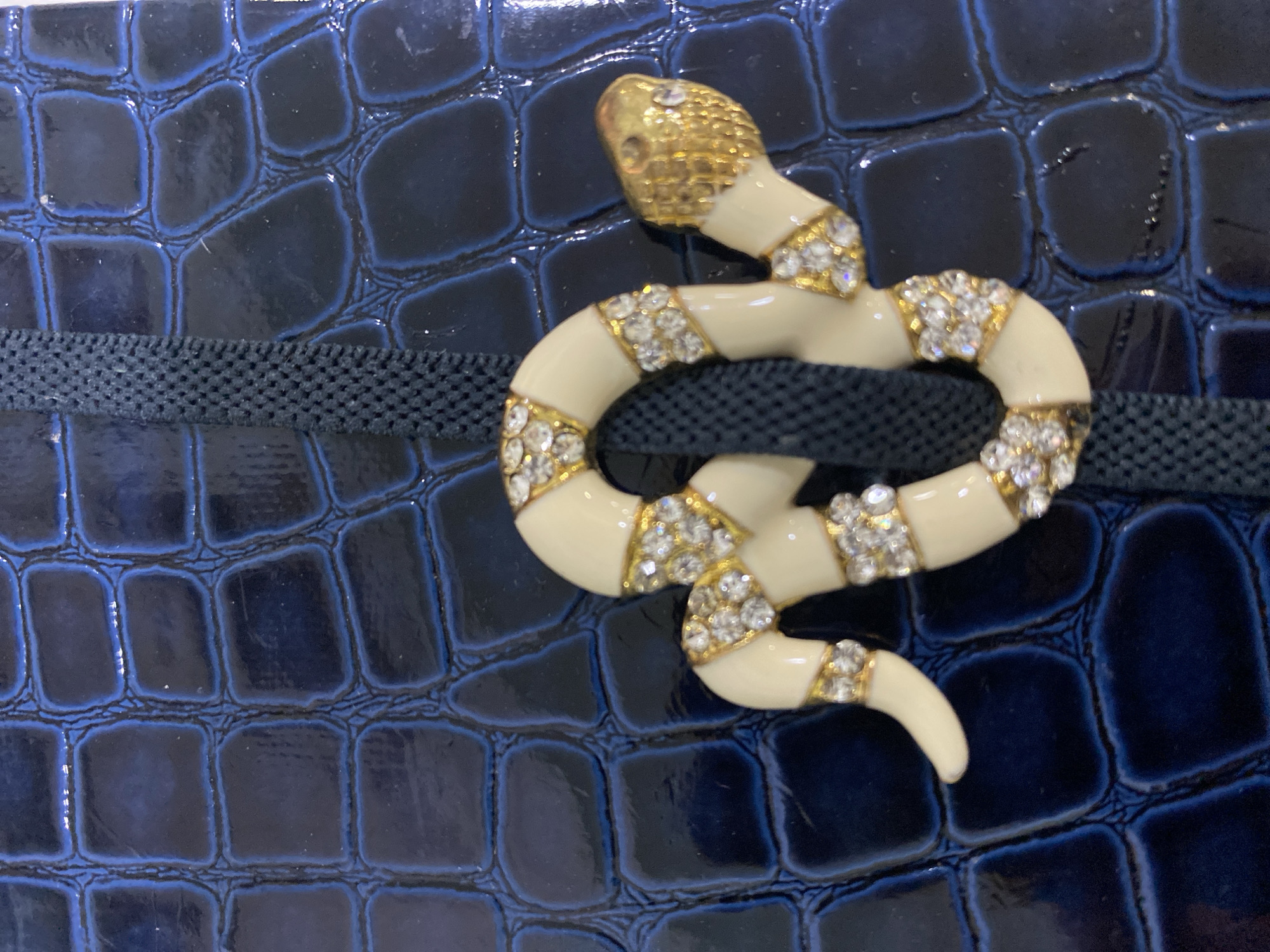 A decorative, coiled snake-shaped object is positioned on a dark blue, glossy surface featuring a simulated reptilian skin texture, located in the city of Rozhny, Ukraine. The snake's body is predominantly cream-white with golden segments, each studded with multiple clear, faceted gemstones. The snake's head is gold-toned and includes small clear gemstones. A dark blue, finely woven strap runs vertically behind the snake, slightly left of the image's center, overlaying the textured background.