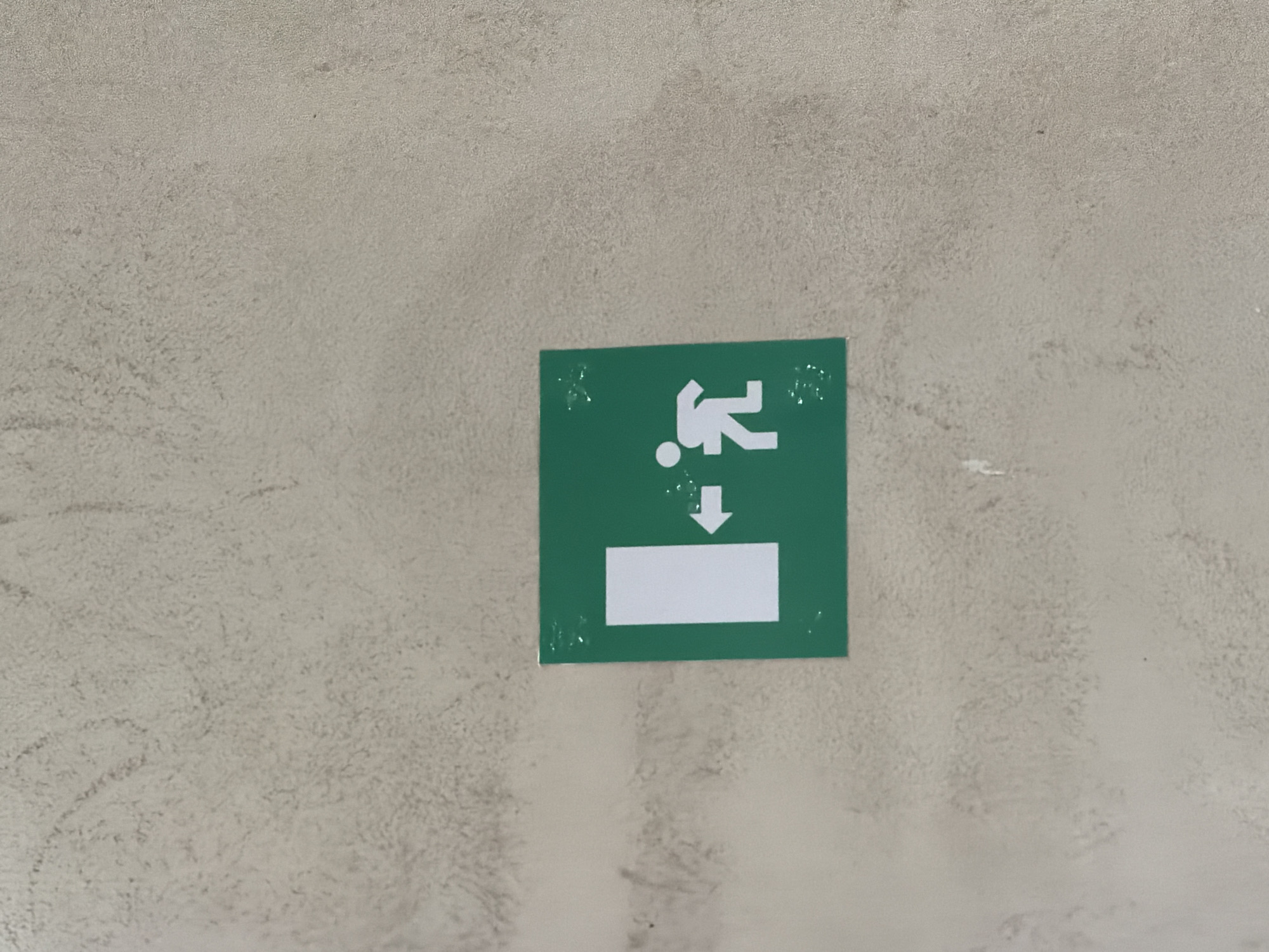 The image shows a green square sign with white pictograms on a textured, light-colored wall. The sign depicts a white rectangle on the left, representing a doorway, with an arrow pointing from the doorway towards a white figure on the right. The figure is stylized and appears to be running or moving away from the doorway. This is a standard exit sign, indicating a place of egress. The wall has an uneven, stucco-like finish with some smudges or dirt marks visible. The lighting suggests an indoor environment. The sign is positioned centrally in the frame. There are no people visible in the image other than the pictogram. The location is specified as Piacenza, Italy.