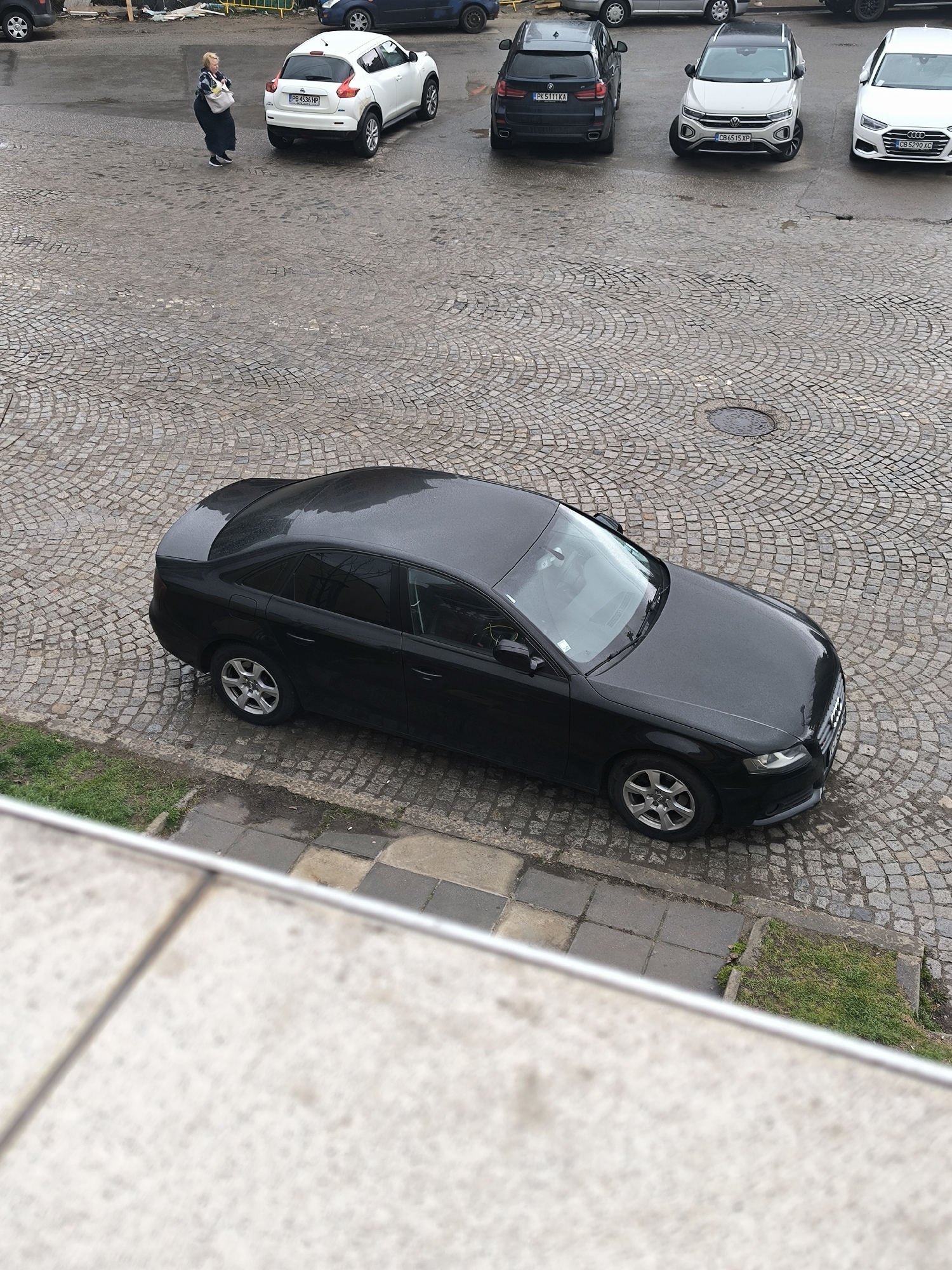 The image presents an elevated view of a cobblestone square in Sofia, Bulgaria. A black Audi sedan is parked prominently in the foreground, occupying the lower-center portion of the frame. The car is adjacent to a concrete curb with patches of grass growing nearby.
In the background, there are several other parked vehicles, including a white Nissan Juke, a dark blue BMW X5, a white Volkswagen T-Roc, and a white Audi A4. A woman wearing a dark coat and carrying a white bag is walking on the square towards the parked Nissan Juke.
The environment appears to be urban, with wet cobblestones suggesting recent or ongoing precipitation. The scene is captured from an elevated position, likely a building or upper-story window.