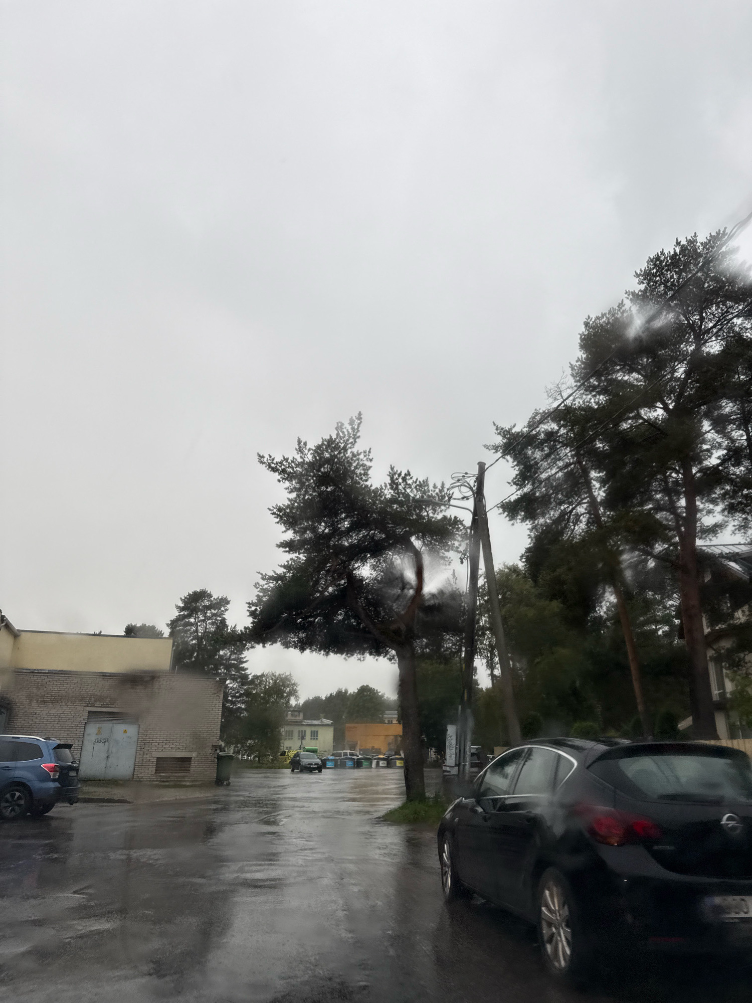 The image depicts a wet asphalt road in an overcast environment, likely during or after rainfall. Several cars are visible: a blue SUV parked on the left, a black Opel hatchback in the foreground, and a few other cars further down the road. The surrounding area includes buildings, trees, and a utility pole. The building on the left is light-colored with a large gray door. A green trash bin is next to it. Further down the road, more buildings and trees are visible. The overall atmosphere is somber due to the weather and the lack of clear activity. The location is likely in Tallinn, Estonia, given the context provided.