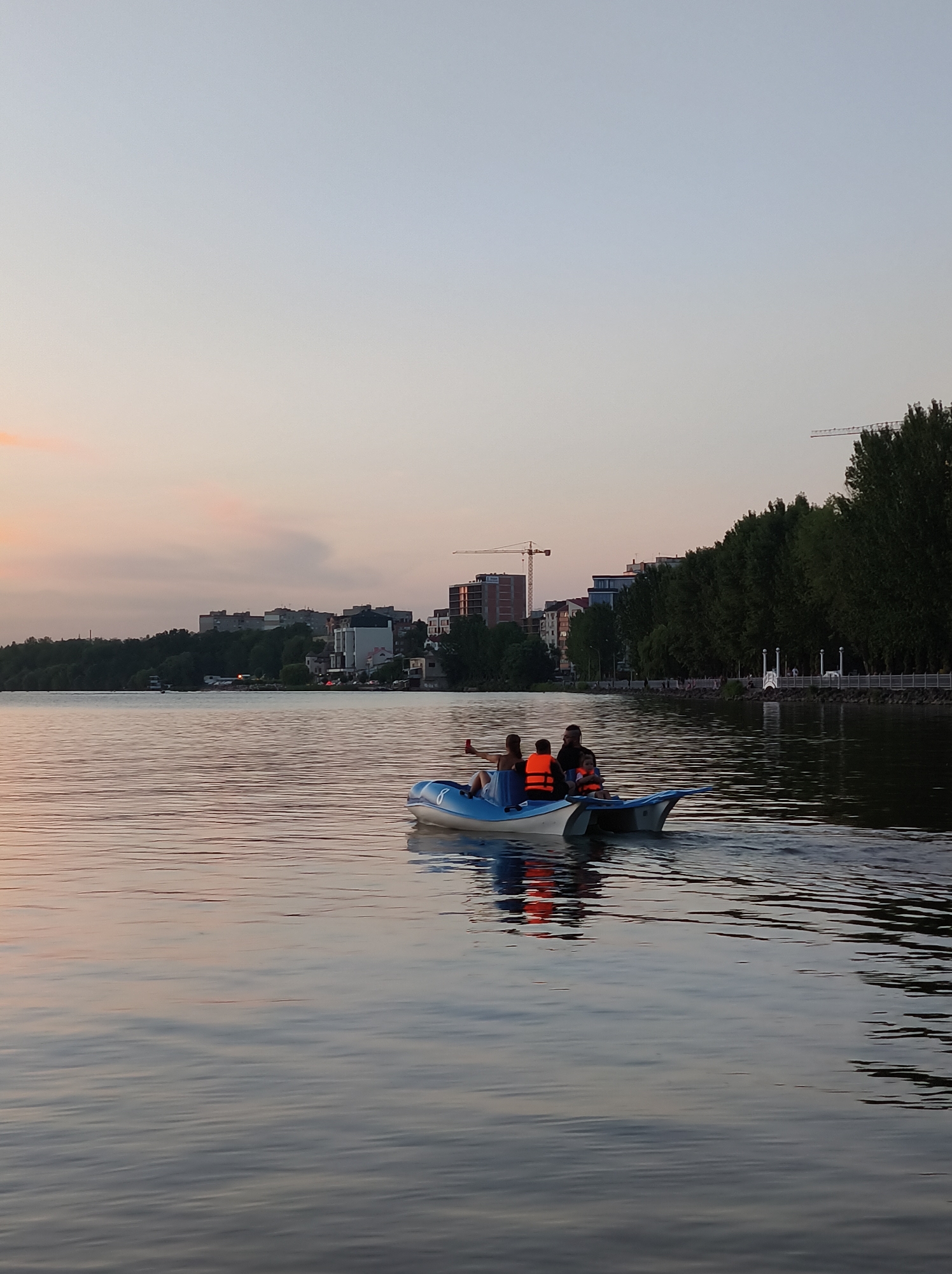 In the city of Ternopil, Ukraine, at dusk, a group of four individuals is enjoying a pedal boat ride on Lake Ternopil. The water reflects the pastel colors of the fading sky and the surrounding cityscape. The pedal boat, a blue and white catamaran-style vessel, is positioned in the middle ground of the image. Two adults and two children are seated in the boat. The person in the front left is taking a selfie with a mobile phone. All occupants are wearing life vests, with three of them in bright orange. The background features a treeline, a promenade with white decorative elements, and a cityscape with several multi-story buildings, including what appears to be a construction crane in the distance. The lighting suggests it is late evening, with a gentle breeze creating ripples on the water's surface.