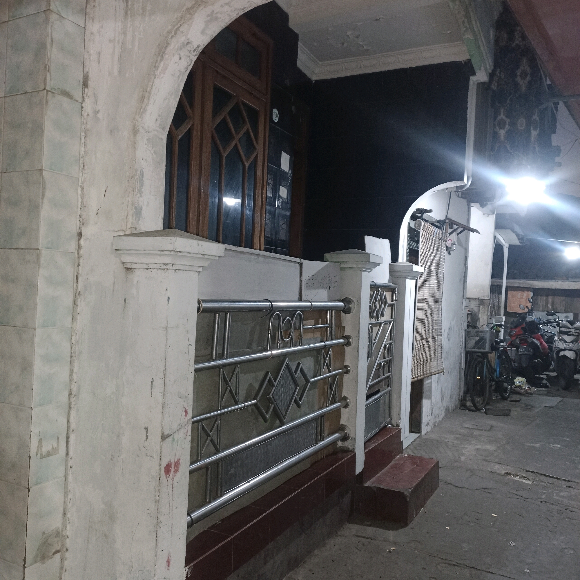 The image captures a dimly lit, outdoor scene at night, possibly in front of a building. In the foreground, a white concrete pillar and part of a metal fence are visible. The fence has a decorative diamond pattern and horizontal bars. To the left of the fence, a section of tiled wall is present, with some tiles appearing light blue or green. Behind the fence, a wooden-framed window with diamond-shaped panes is set into a dark-tiled wall.
Further back, a bamboo-slatted blind hangs on the wall. The ground is paved with a rough, gray surface. On the right side of the image, several motorcycles are parked, suggesting a residential or commercial area. One motorcycle is red, and others are partially visible. A bicycle is also parked near the motorcycles. The overall impression is of a modest, somewhat aged facade. The scene is illuminated by artificial light sources, including a bright light near the top right that creates a lens flare effect. There are no people or animals clearly depicted in the image. The location is identified as a specific alley in the city of Surabaya, Indonesia, known for its residential character and evening activity.