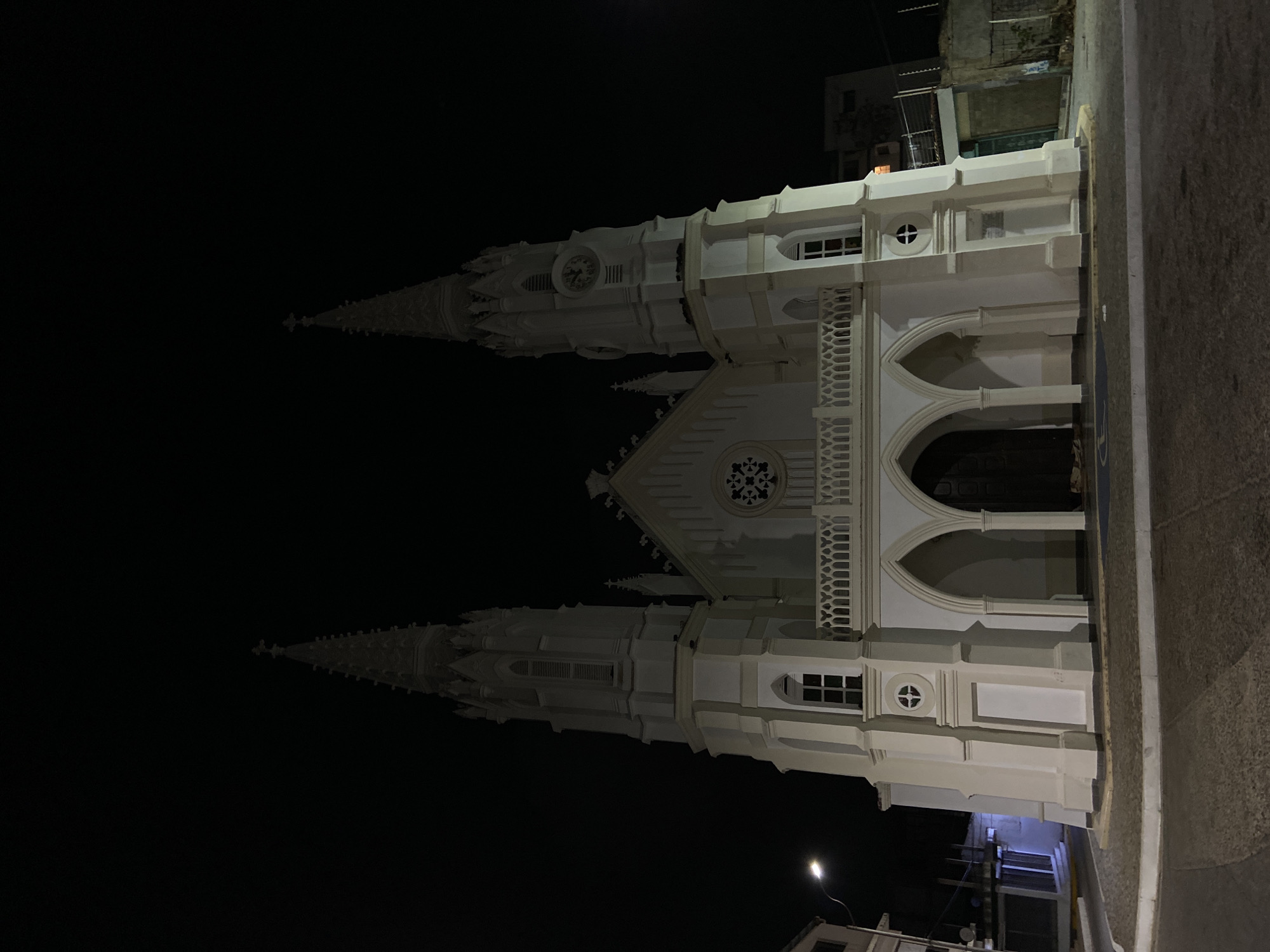 The image depicts the Church of San Juan Evangelista, also known as the Iglesia de Juan Griego, in Juan Griego, Venezuela, at night. The church is illuminated and stands out against the dark sky. The architecture is characterized by two prominent towers with spires, Gothic-style arches, and white facade. The image is taken from street level, with a portion of the adjacent road visible.