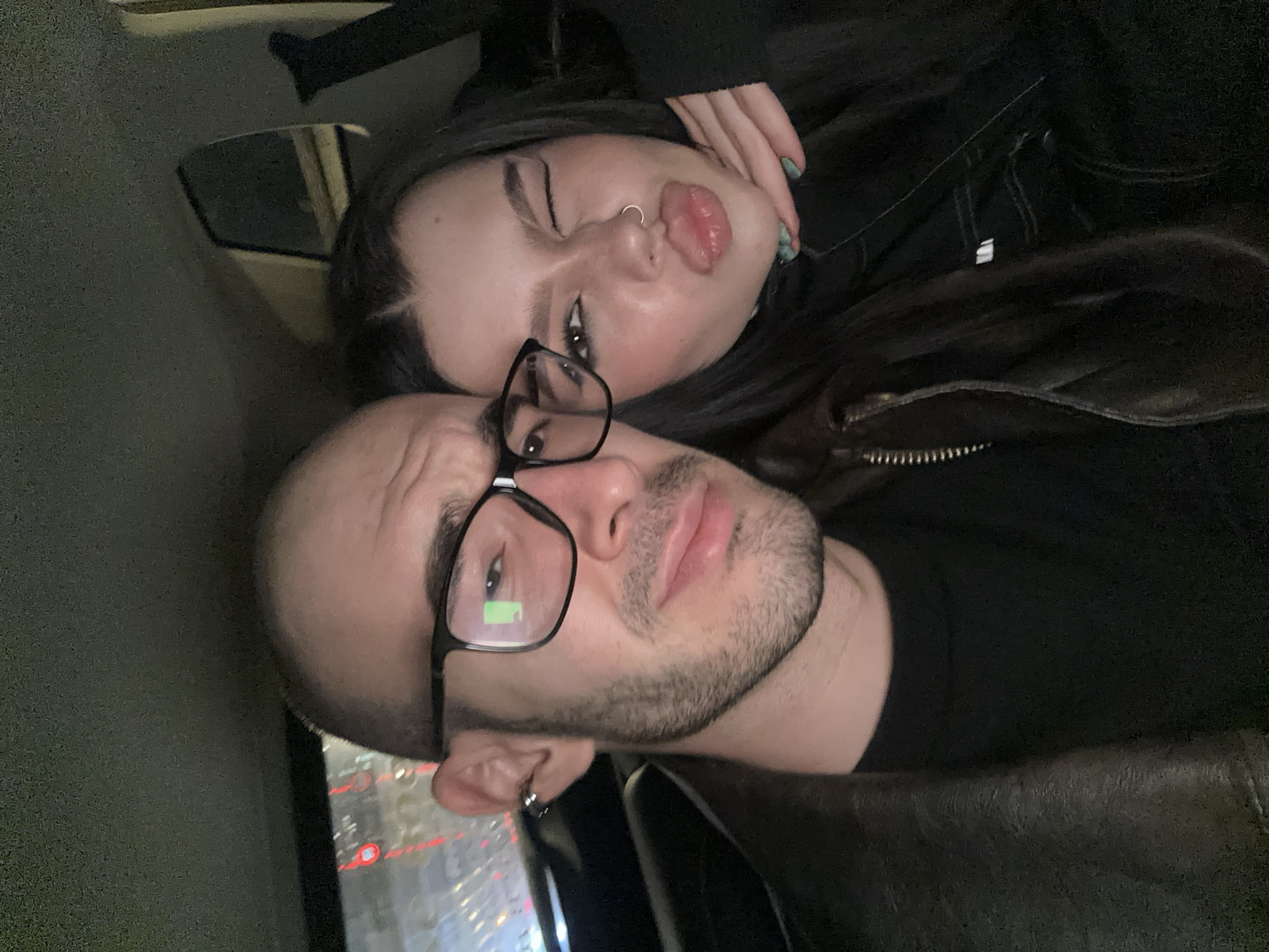 Two people are posed closely together for a selfie. The man is bald, with a beard, and wearing glasses. The woman has long dark hair and is wearing a nose ring. They appear to be inside a car, with reflections of city lights visible through the window. The location is likely Moscow, Russia, based on the appearance of the buildings and streetlights.