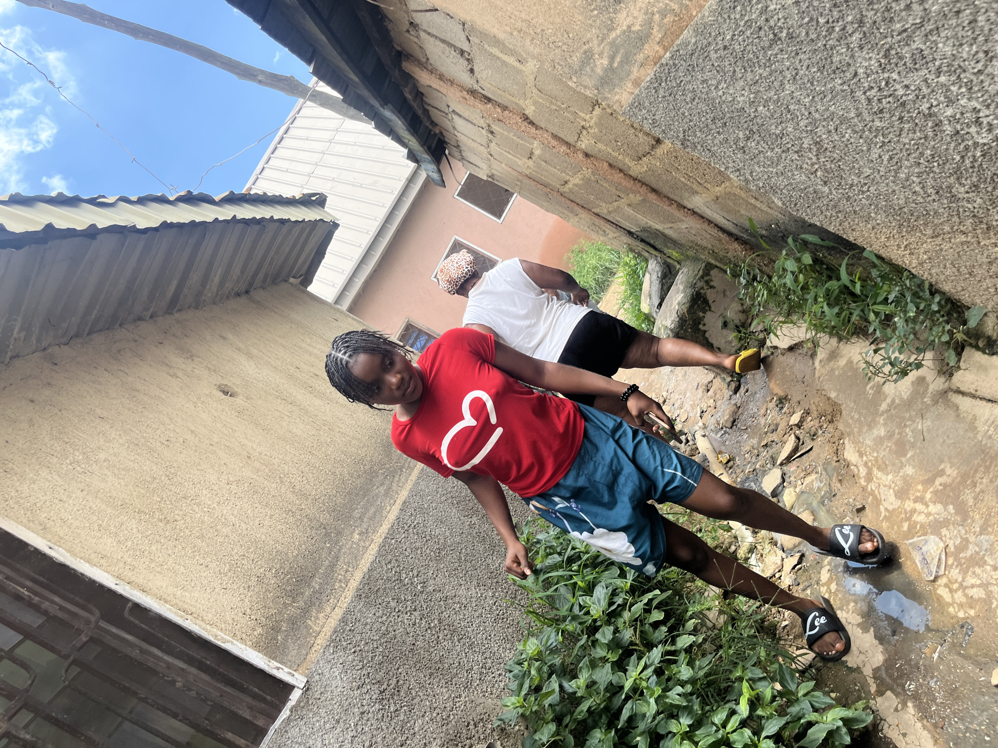 The image shows a young woman in a red t-shirt with a white heart emblem and blue shorts walking on a dirt path. She is wearing black sandals. Behind her, another person in a white t-shirt and black shorts, wearing a patterned headscarf, walks away from the camera. The scene appears to be in a narrow, possibly residential alleyway in Jos, Nigeria, with rough stone and concrete walls on either side. There is some green vegetation growing in patches along the path and walls. The ground is uneven with puddles of water, suggesting recent rain or poor drainage. The background reveals buildings with corrugated metal roofs and a bright blue sky with scattered clouds.