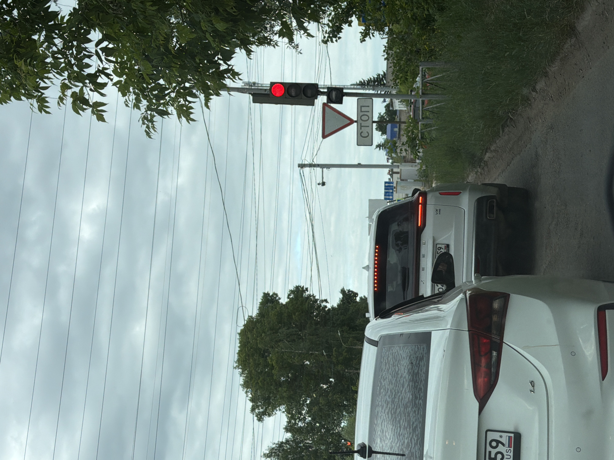 The image depicts a traffic scene on a cloudy day in Chaikovsky, Russia. In the foreground, the rear of a white sedan is visible, facing away from the camera. The car's license plate displays "59 RUS." Further ahead, a white SUV is stopped in traffic, also facing away. Behind the SUV, other vehicles and buildings are partially visible. To the right of the road, a traffic light is red, and a stop sign with the Cyrillic word "СТОП" is displayed below a yield sign. The environment includes trees and power lines. The road surface appears to be asphalt.