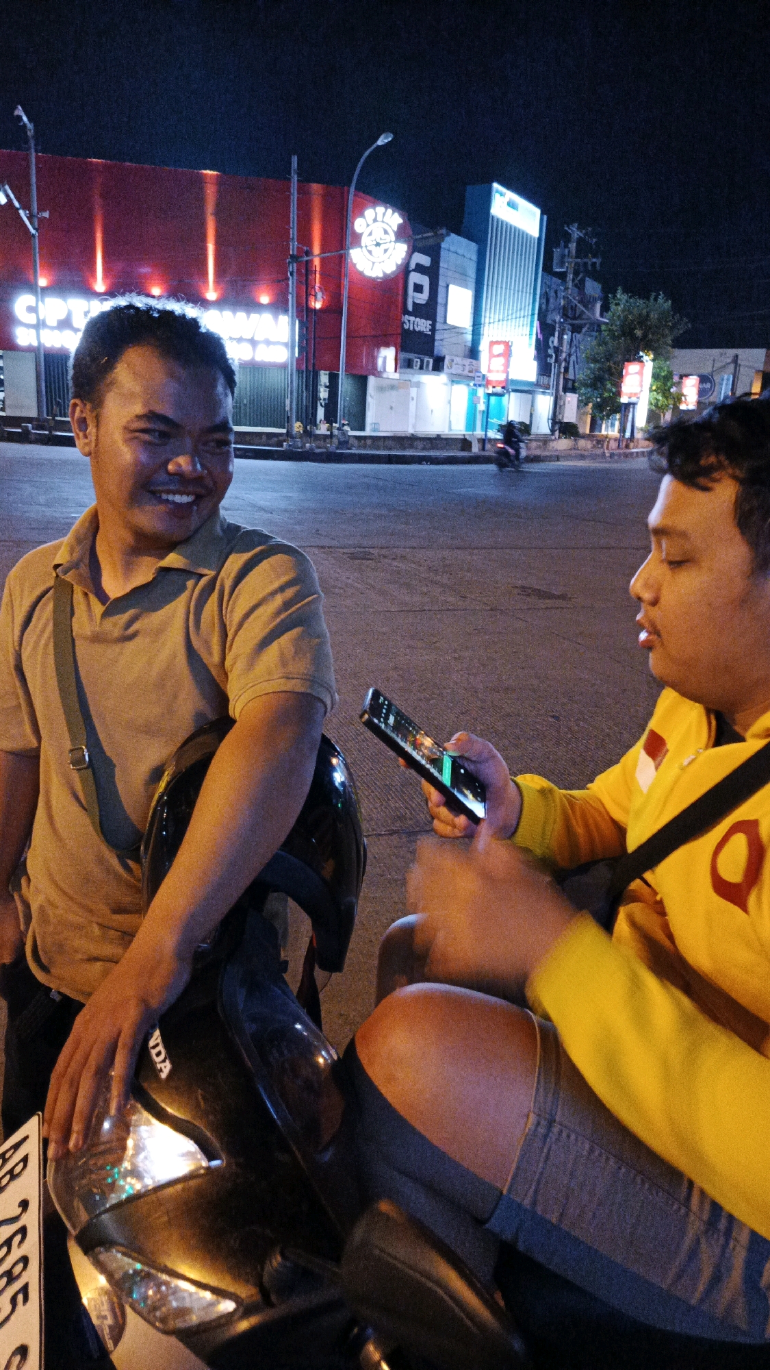This image depicts a nighttime scene on a street in Surakarta, Indonesia. Two men are visible, seated on a motorcycle. The man on the left, wearing a light brown polo shirt, is smiling and looking to his left. He has a dark helmet in front of him. The man on the right, wearing a yellow hooded jacket, is holding a smartphone and looking at it. He is wearing grey shorts. The background shows a street with buildings and lights. One building prominently features a large red facade with illuminated signage and a circular logo. Another building is tall and modern, illuminated with blue lights. Traffic lights and street lamps are also visible. The street surface is asphalt, and there are some trees and foliage in the distance. A motorcycle license plate, partially visible on the left, reads "AB 2685 S".