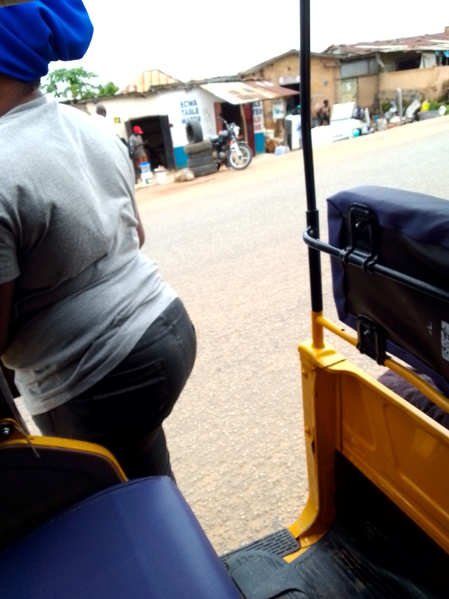 A person wearing a blue head covering and a grey t-shirt is seen from behind, standing next to a yellow and blue vehicle, likely a commercial tricycle, in a street scene in Jos, Nigeria. The person is wearing dark pants and appears to be either boarding or disembarking the vehicle. In the background, there are buildings, a motorcycle, and stacked tires, suggesting a commercial area. The ground is paved with asphalt and gravel. The overall impression is of a busy street in an urban setting.