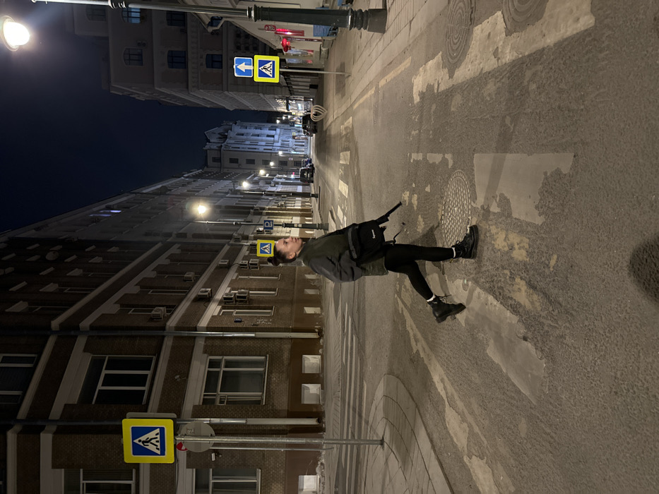 A nighttime street scene on Petrovskie Linii Street in Moscow, Russia, depicts an adult female pedestrian walking from right to left across a marked crosswalk. She wears a dark jacket, black leggings, and black combat boots with white socks visible. A black shoulder bag with a white "Supreme" logo is worn across her body. In her right hand, she carries a dark, long object resembling a rifle or shotgun, oriented downwards and slightly forward. The street is asphalt with visible, partially worn white pedestrian crossing stripes and a circular manhole cover. Multi-story brick and lighter-colored buildings line both sides of the street, featuring windows and external air conditioning units. The scene is illuminated by multiple streetlights. Road signs include blue pedestrian crossing signs (white figure on blue square) on both sides of the street, and a blue upward arrow sign (one-way street) on the right. Several parked cars are visible further down the street. A decorative heart-shaped structure stands on the sidewalk to the right.