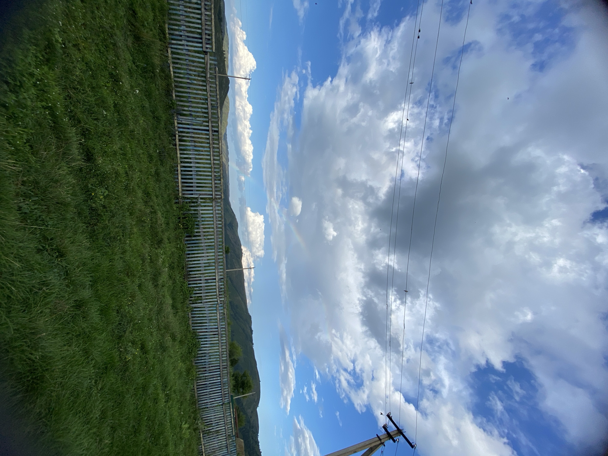 The image depicts a rural landscape under a partly cloudy sky. In the foreground, lush green grass covers the ground. A weathered, light blue wooden fence runs horizontally across the middle ground. Beyond the fence, rolling green hills stretch towards the horizon, with a faint rainbow visible in the distance. A utility pole with wires is visible on the right side of the image, and a few birds are perched on the wires and flying in the sky. The scene appears to be in Maloarslangulovo, Russia, suggested by the rural setting and style of the fence and utility pole.