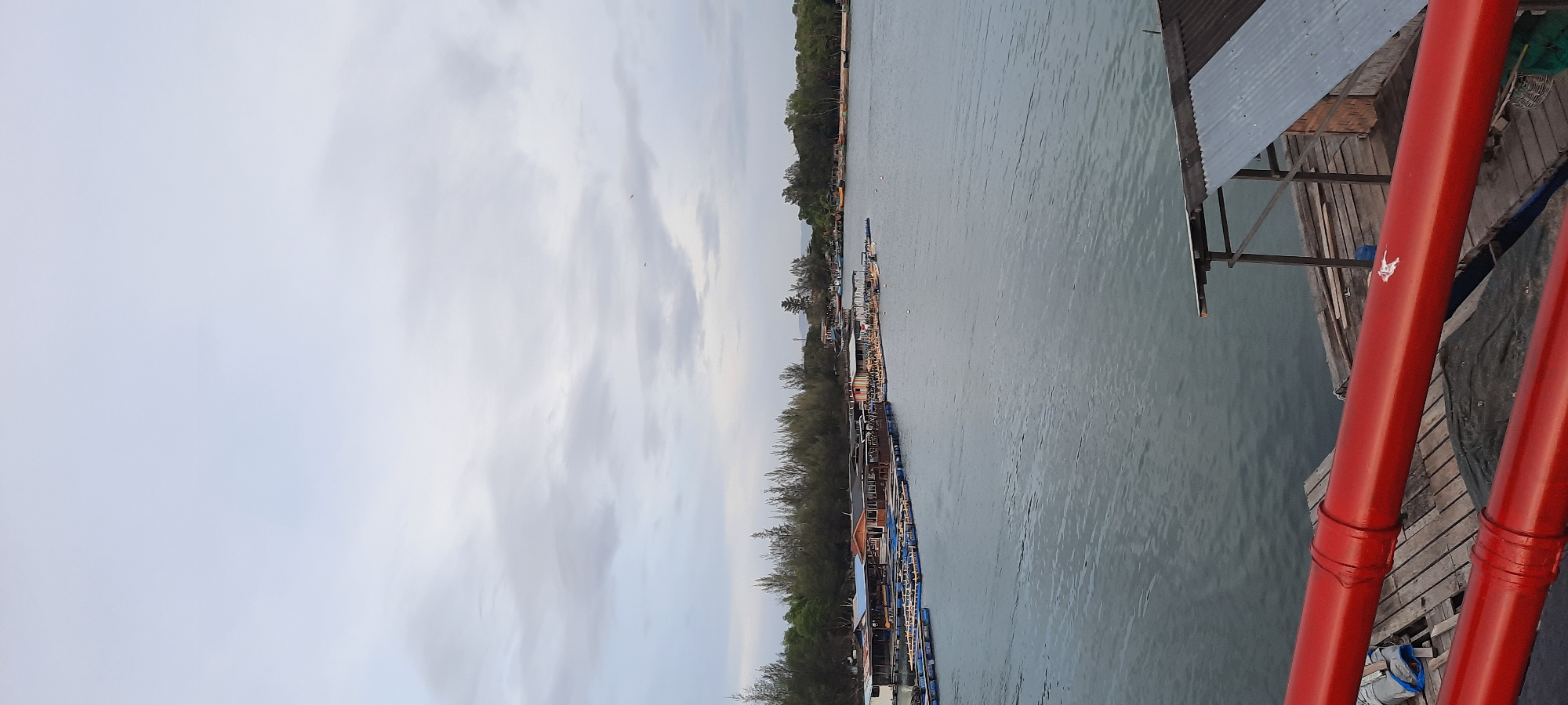 The image shows a waterfront scene, likely in Banda Aceh, Indonesia, characterized by a grey, overcast sky. The water is calm and grey, reflecting the cloudy sky. In the mid-ground, a collection of floating structures, possibly fish farms or restaurants, are visible. These structures are connected and extend along the shoreline, which is lined with dense green trees. A few small boats are docked or moored near these floating platforms. In the foreground, there are two bright red pipes, which appear to be part of a pier or some sort of industrial structure. The pipes are positioned horizontally across the frame, with wooden planks and a small, corrugated metal-roofed shed visible beneath them. The overall impression is one of a working waterfront, possibly a tourist area given the presence of what looks like dining platforms.