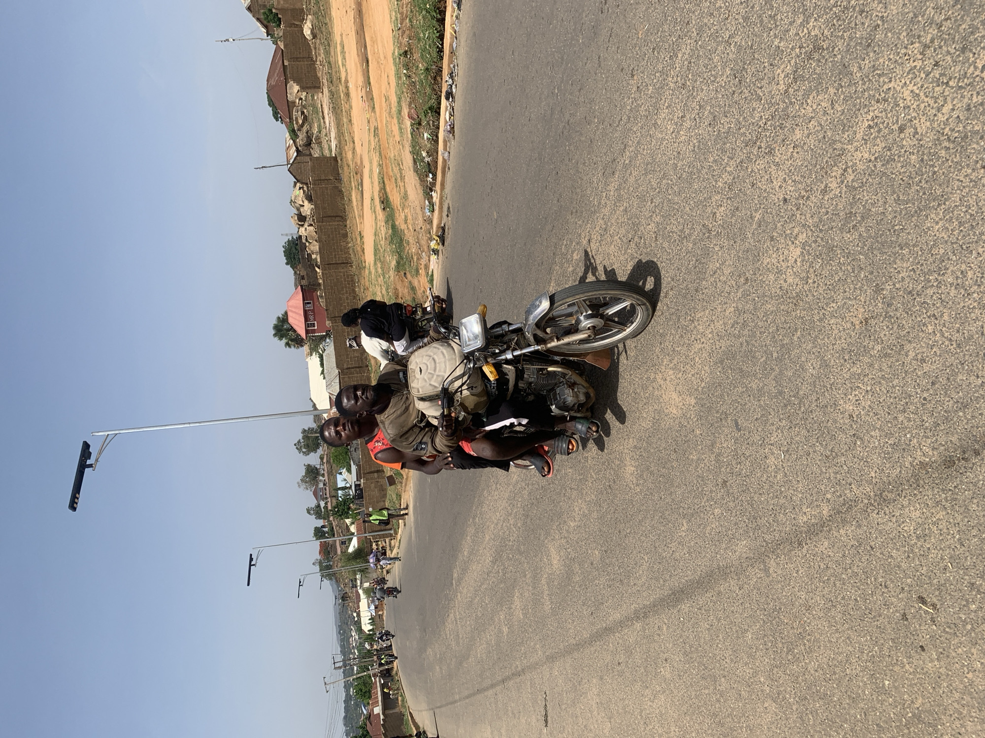 The image shows two men riding a motorcycle on a paved road in Gura Top, Nigeria. The man in the front is wearing an orange shirt, while the man behind him is wearing a dark-colored shirt. The motorcycle is facing right with the front tire pointed toward the viewer. The buildings and vegetation are visible on the left side of the road. The sky is clear. There are street lights along the road. Other motorcycles can be seen in the distance.