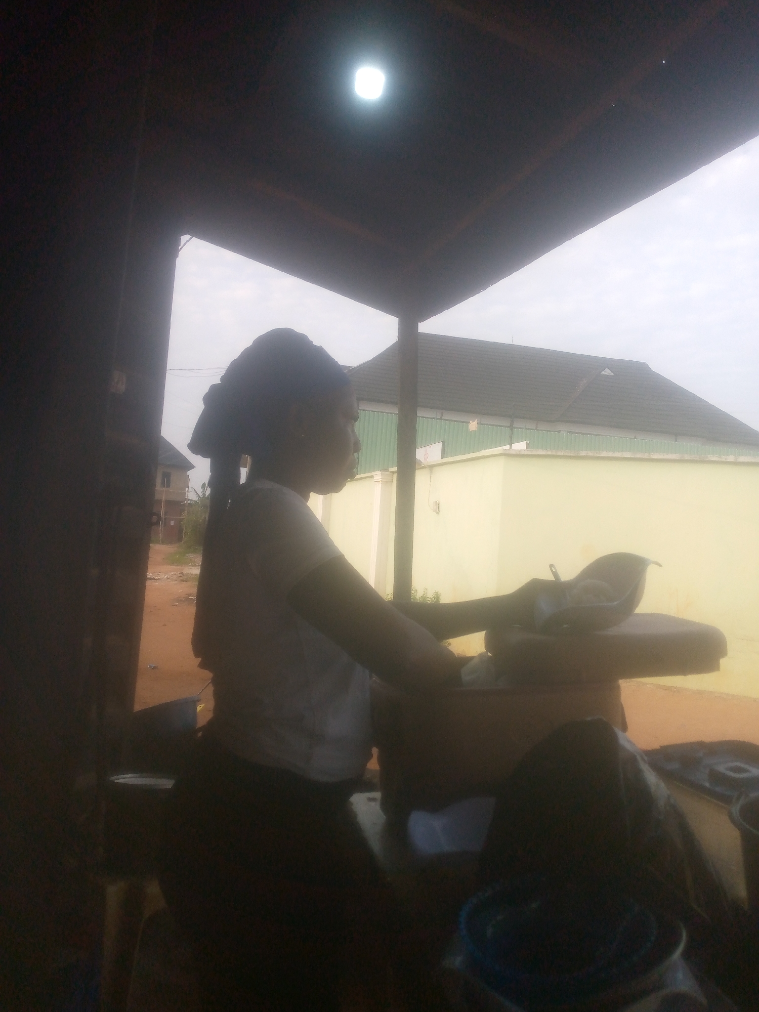 A woman stands in a dimly lit outdoor workspace under a makeshift roof, possibly a stall or shed. She is wearing a white T-shirt and dark pants. She is working at a station that contains some metal objects and an ironing board on top of which there is an object resembling a baby seat, which has something inside it. Outside the workspace is an open area with a beige wall and a roofed house with brown shingles. The image is taken in Unknown, Unknown.