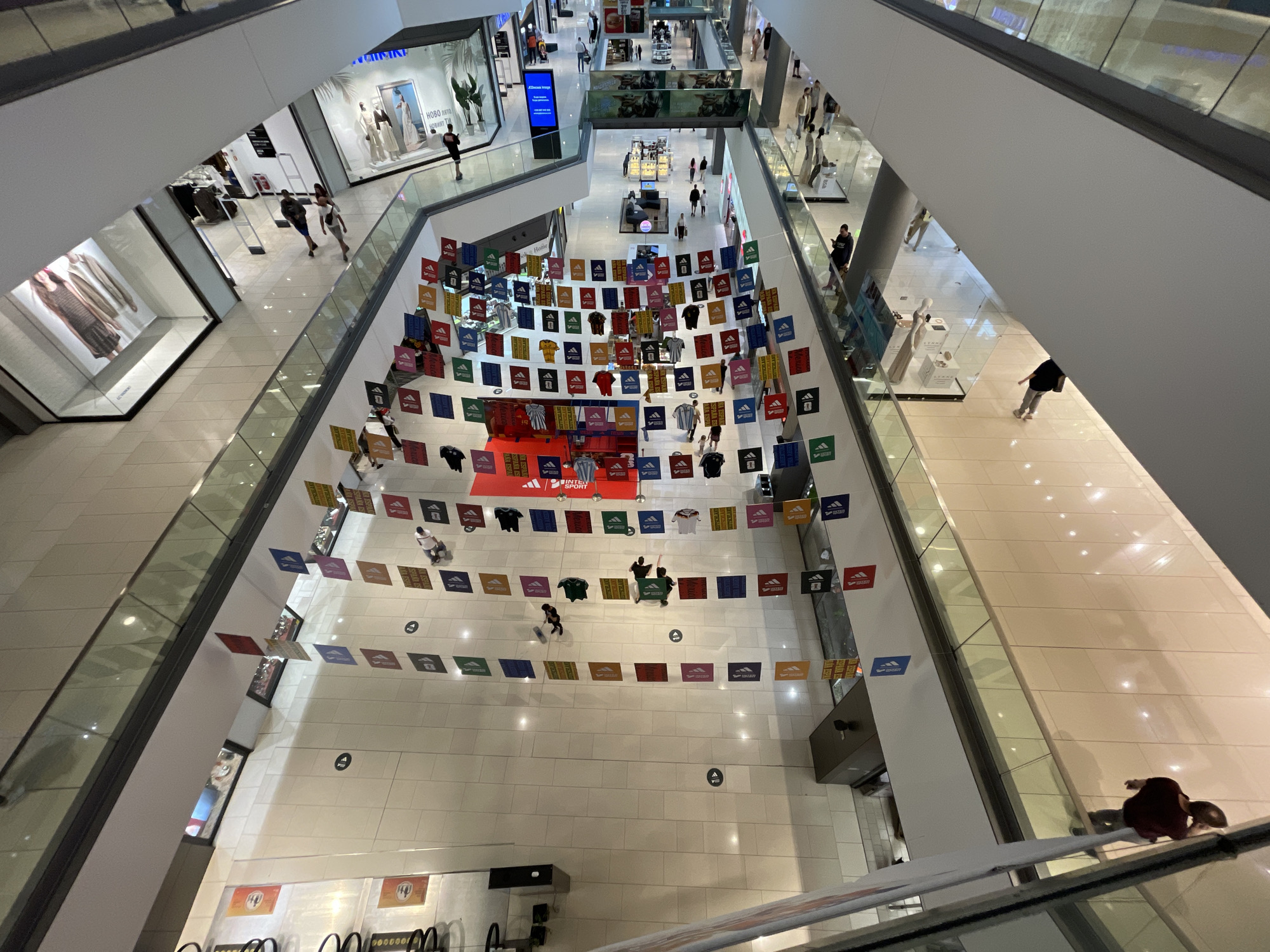 This is an aerial view of the interior of a multi-level shopping mall. The mall appears to be modern with white tiled floors, glass railings, and large display windows for shops. In the foreground, there is a light-colored escalator and a portion of a darker escalator. Several levels of the mall are visible, with escalators connecting them.
A prominent display of Adidas branded merchandise is hung across one of the central open areas. This display consists of numerous colored flags or banners, many featuring the Adidas logo and some displaying Adidas t-shirts and soccer jerseys. There are also some larger red banners with Adidas branding.
People are visible walking on various levels of the mall, browsing in stores, and using the escalators. Some individuals are walking alone, while others are in small groups. The environment appears clean and well-lit, typical of a retail shopping center. The specific location is identified as The Mall in Sofia, Bulgaria, given the presence of Adidas merchandise and the mall's architecture.