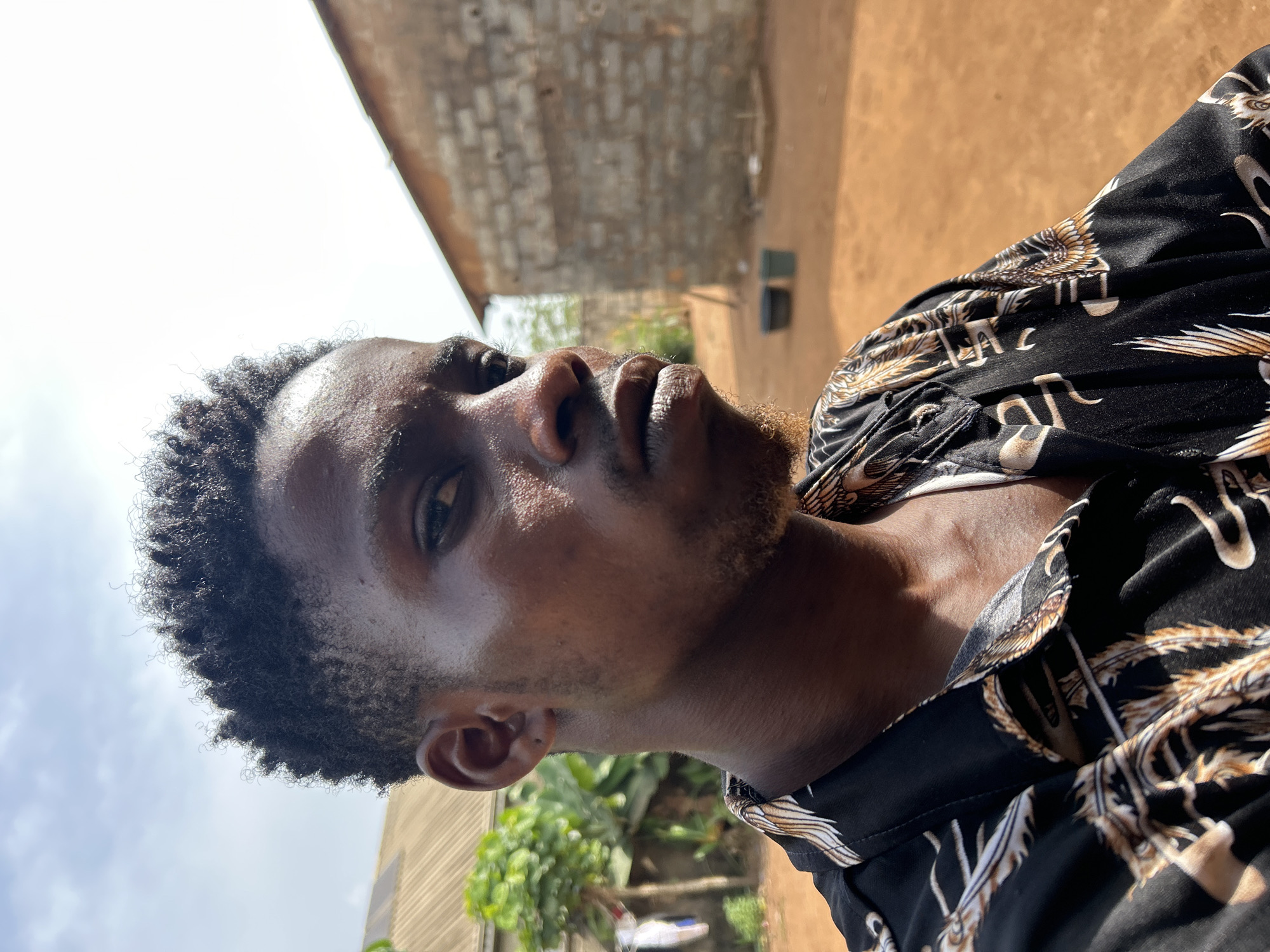 A medium, eye-level shot shows a Black man with dark, curly hair and facial hair, wearing a patterned black and gold shirt. He's looking upward and slightly to the left, with a serious expression. His skin appears to be slightly damp, possibly from sweat. The background consists of a building constructed from light grey bricks, along with some greenery and what appears to be a dirt or clay ground. The sky is visible in the upper left corner, appearing partially cloudy. The scene is outdoors, likely during daylight hours in Unknown, Unknown.