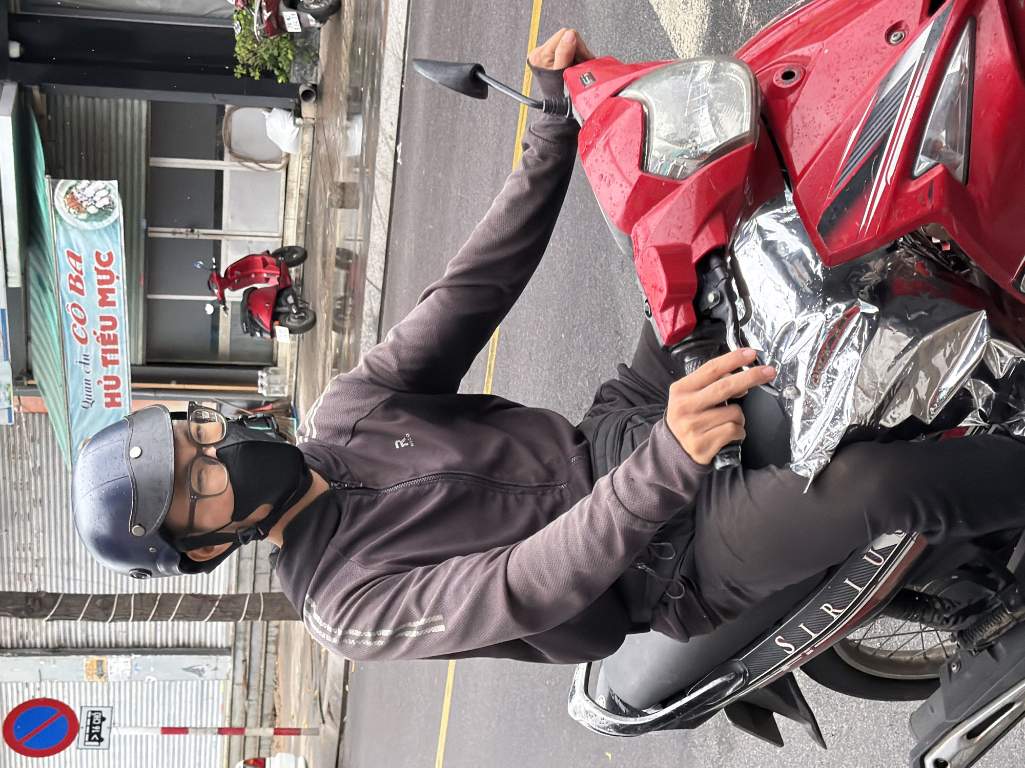 A person wearing a dark gray hooded jacket, black pants, black mask, and a blue helmet rides a red scooter on a wet street in Nha Trang, Vietnam. The person is wearing glasses and the scooter has a silver emergency blanket covering part of its front. The background shows a closed storefront with a sign that reads "An Co Ba Hu Tieu Muc" and a red parked scooter. A traffic sign indicating "No Entry" and "16 Cho" is visible on the left side of the image. The street is wet and reflective, suggesting it may have recently rained.