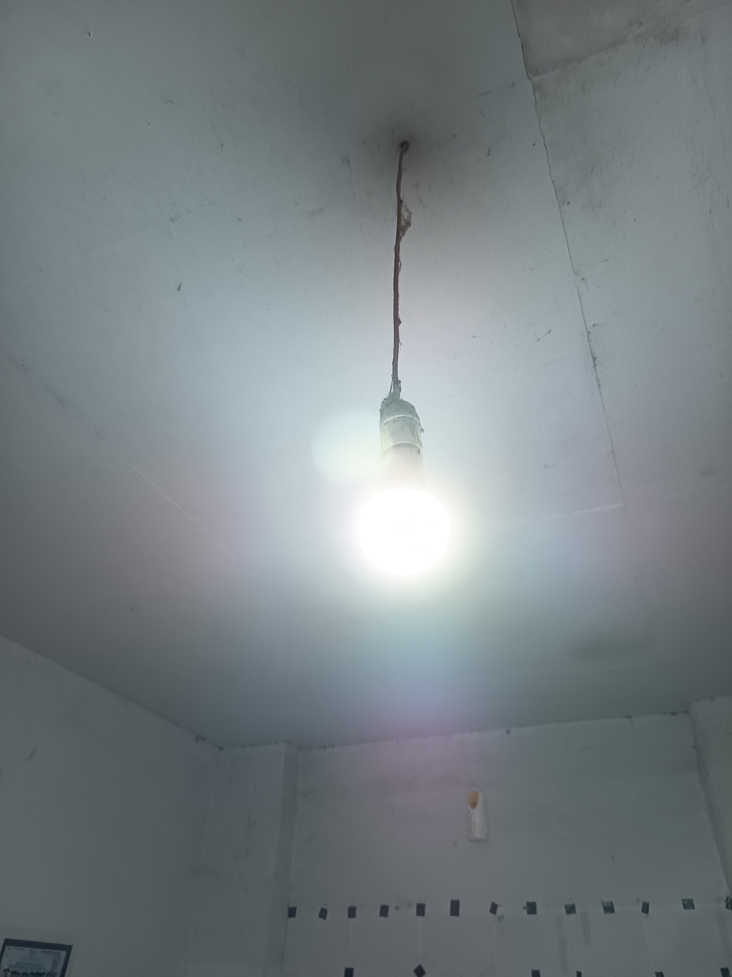 A lit light bulb hangs from a cord attached to a white ceiling in an unfinished room. The bulb is bright and casts a halo of light. The ceiling has some cracks and marks, and appears to be made of plasterboard panels. The walls are also white and appear to be unfinished. In the lower left corner, a framed object is partially visible on the wall. In the background, along the wall, there are a series of dark square markings. There are no people in the image. The environment suggests a construction or renovation site. This scene is not in Krampilan, Indonesia.