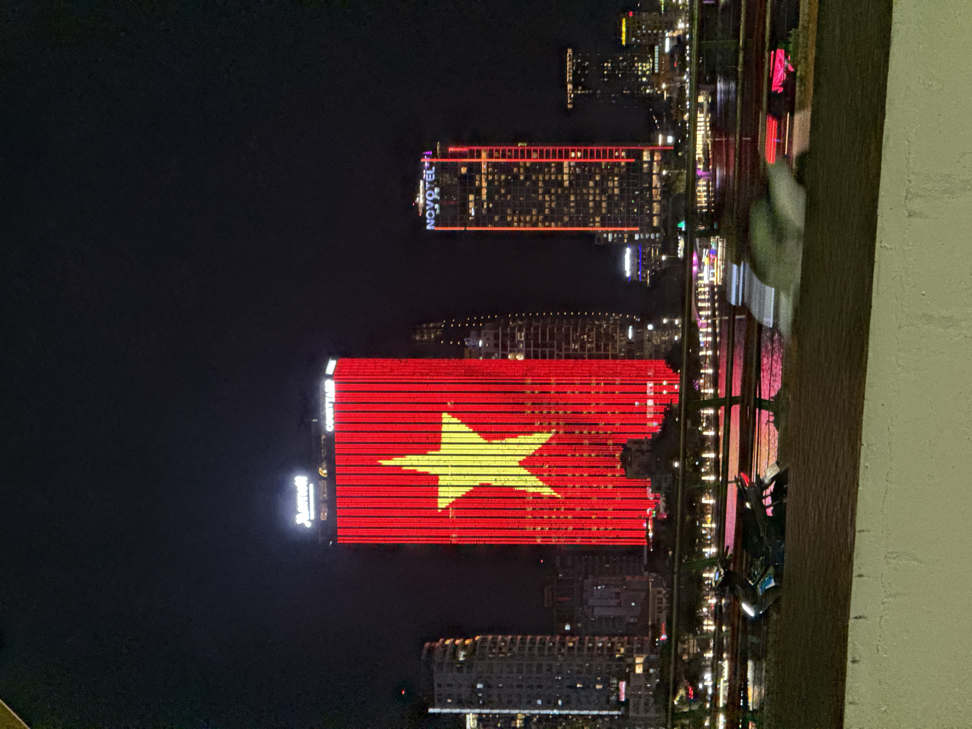 The image shows a nighttime cityscape with several tall buildings illuminated by lights. The most prominent building is lit up with the Vietnamese flag, featuring a red background and a large yellow star. To the right, another building displays the "NOVOTEL" logo in illuminated letters. In the foreground, a body of water reflects the lights of the city, and a bridge can be seen spanning it. A motorcycle is parked on the left side of the foreground. The city appears to be Đà Nẵng, Vietnam, based on the presence of the Vietnamese flag displayed on a prominent building, a common patriotic display in Vietnamese cities. The overall scene depicts a vibrant urban environment at night, with architectural landmarks and the visual dynamism of city lights.