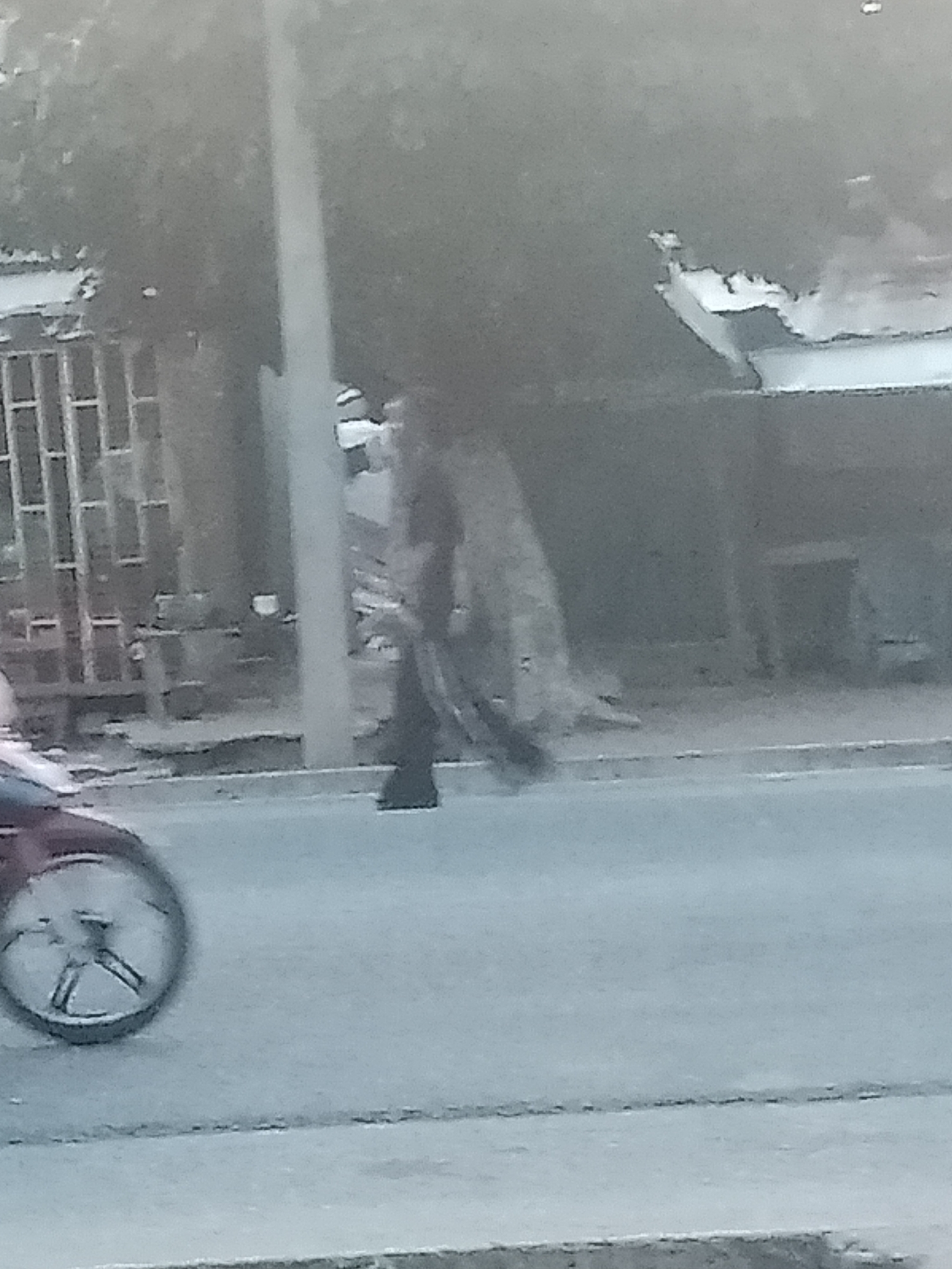 A person wearing dark clothing walks along a paved road in Jalingo, Nigeria. The person is in the middle ground of the image, positioned slightly to the left of center. A portion of a dark-colored motorcycle is visible in the lower left corner, with its wheel clearly in frame. In the background, a building with an open structure and a barred window is partially visible. A tall, slender pole stands vertically to the left of the walking person. The overall scene appears to be an outdoor urban environment, with the road dominating the foreground.