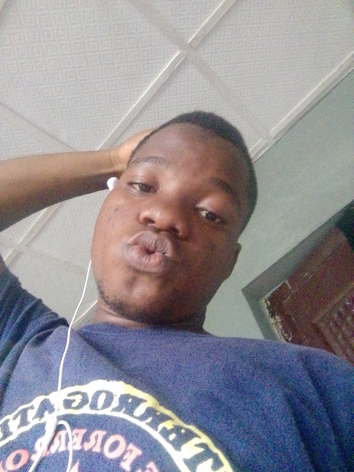 A young Black man is captured in a close-up selfie, his head tilted slightly and his hand resting on the back of his head. He is wearing a dark blue t-shirt with a yellow and pink circular graphic on the front, and white earbuds are visible. His lips are puckered in a kissing gesture. The background shows a textured, light-colored ceiling and a portion of a wall with a dark brown door or cabinet. The image appears to have been taken indoors, possibly in a residential setting in Ipetumodu, Nigeria.