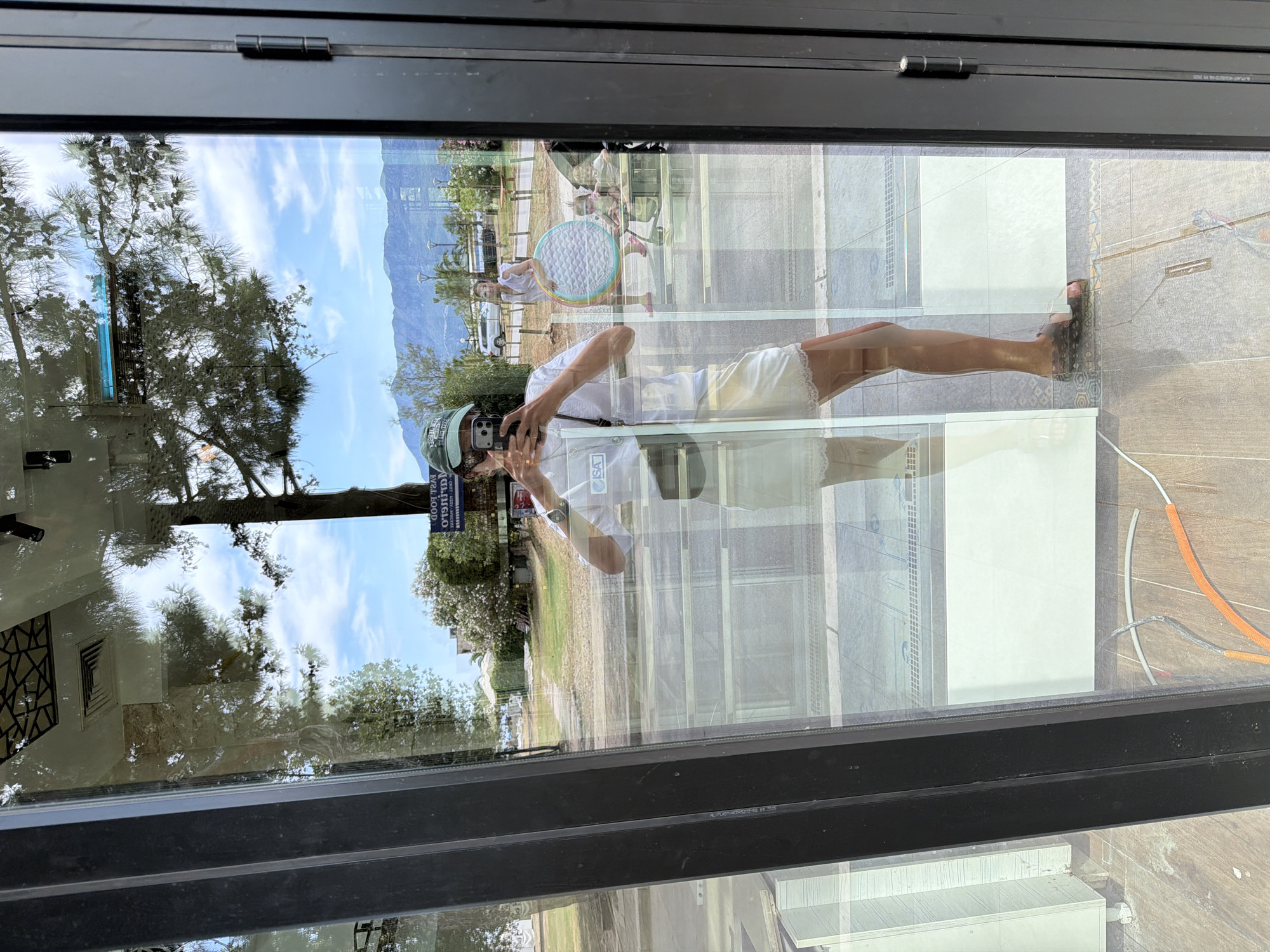 A person, seen in a reflection in a glass storefront, is taking a selfie with their smartphone. The person is wearing a green cap, sunglasses, a white t-shirt, white shorts with lace trim, and dark sandals. They have a black strap across their chest. In the reflection, a woman is visible in the background holding a colorful inflatable toy, and two young children are seated in a stroller or high chair. The setting appears to be a street in Budva, Montenegro, with palm trees, buildings, and mountains visible in the distance. The storefront has black framing and the floor inside is tiled. Electrical wires are visible on the floor in the foreground.