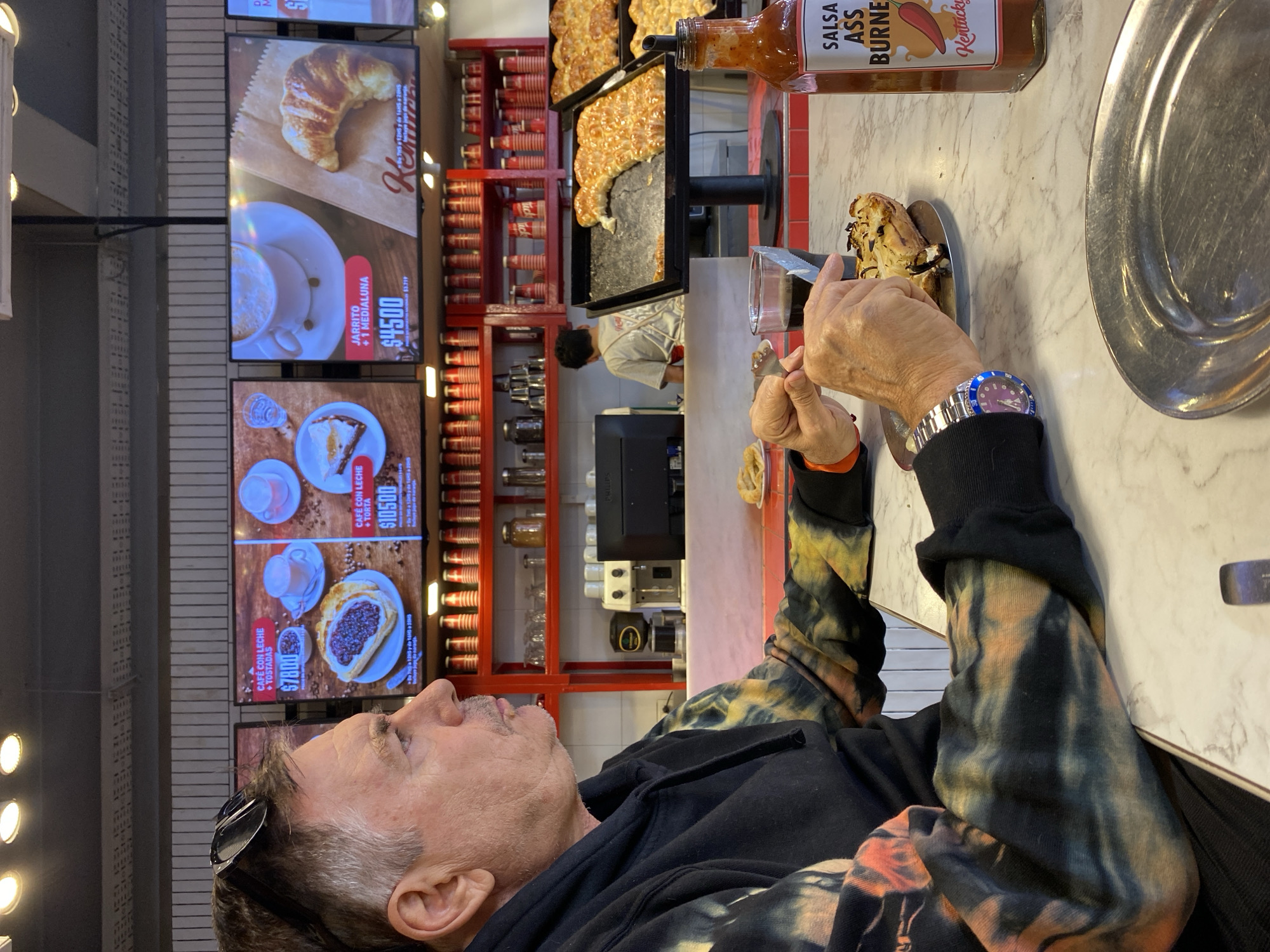 This media file depicts an indoor scene at a casual eatery, likely a café or food court in Buenos Aires, Argentina. The setting is brightly lit, suggesting daytime.
In the foreground, a man with graying hair and a mustache is seated at a white marble-patterned counter. He wears sunglasses perched on his head and a distinctive black, orange, and blue/green tie-dye hoodie. His hands are visible, holding a fork and knife, as he cuts into a pastry or similar food item on a small plate. A blue and silver watch is worn on his left wrist. He appears to be looking up towards the menu screens.
Above and behind the man are multiple digital menu screens displaying food items and prices. Visible text includes "JARRITO + 1 MEDIALUNA $4500", "CAFÉ CON LECHE + TORTA $10500", and "CAFÉ CON LECHE + TOSTADAS $7800". These items and prices, particularly "medialuna" and the amount in Pesos, align with the provided location context of Buenos Aires.
On the counter to the right, a bottle of hot sauce labeled "SALSA ASS BURNER" with the brand "Kentucky" is prominently displayed. Further back, large trays of baked goods, resembling pizza or focaccia, are visible on display racks. Red shelving units are stacked with numerous disposable cups. A "PHILIPS" brand coffee machine is also visible behind the counter, where a partially obscured staff member in an apron can be seen.
The activity centers around the man enjoying his meal in a casual dining environment. The scene captures a moment of everyday life in an urban eatery.