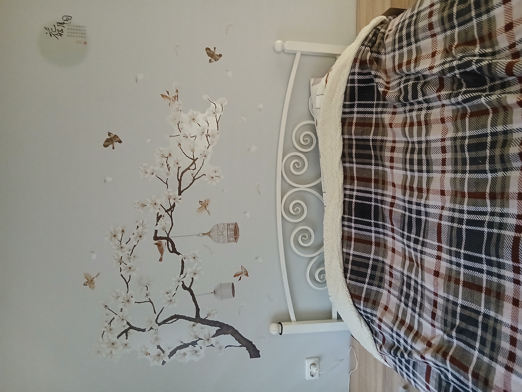 This is a photograph of a bedroom. The focal point is a bed with a white metal headboard, covered by a plaid blanket and a cream-colored sherpa throw. The wall behind the bed is painted a light blue and is adorned with a decal of a flowering tree with birds and decorative birdcages. In the upper right corner of the wall, there is a circular artwork with Chinese calligraphy. To the left of the bed, an electrical outlet is visible with a cord plugged into it. The floor is made of light-colored laminate. The photograph does not provide any information about the city of Mykhailivka, Ukraine, or any people in the scene.