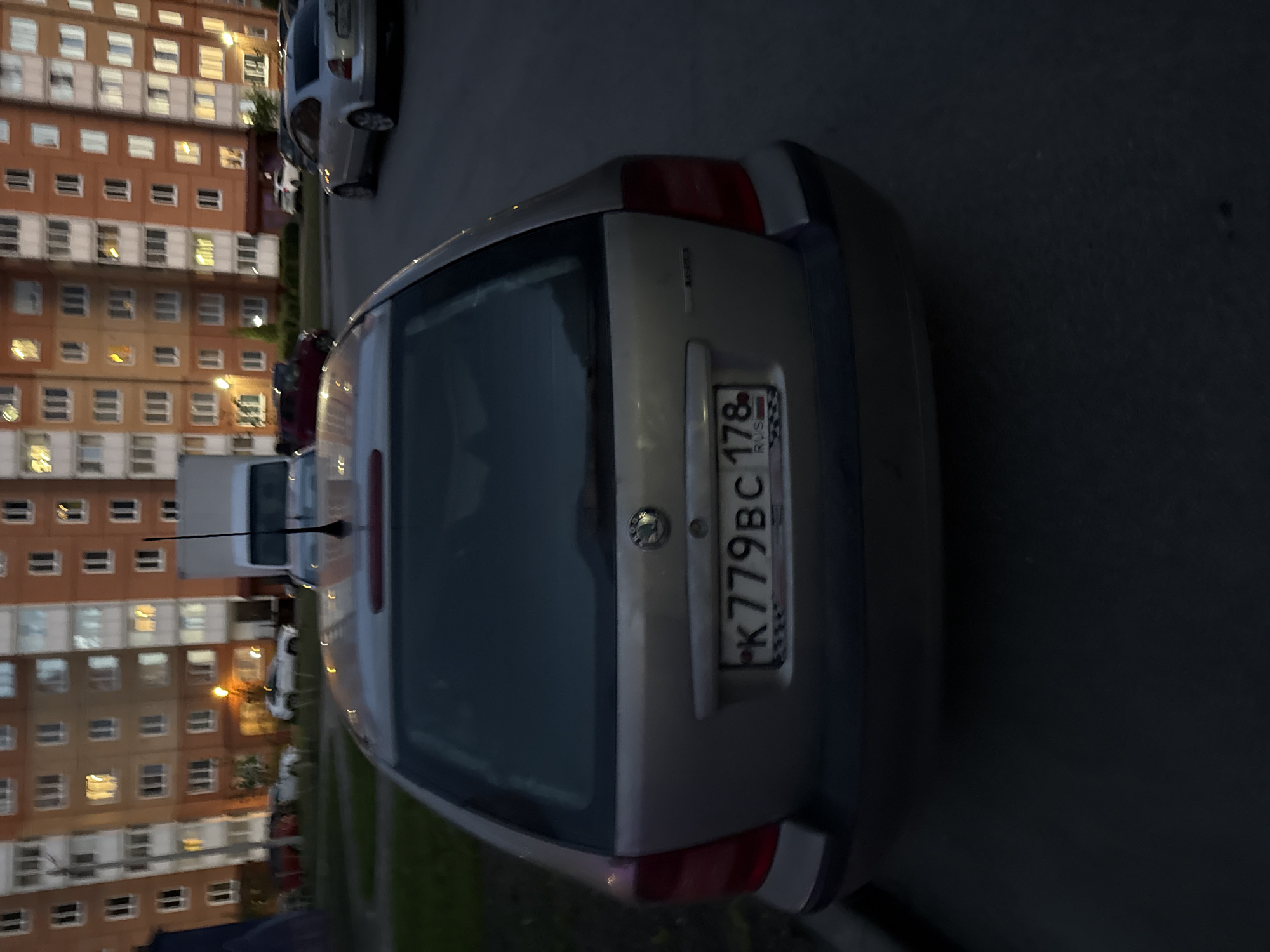 A silver station wagon is parked in front of a tall apartment building. The car's license plate reads "K779BC 178 RUS". The building has multiple floors with windows illuminated by interior lights, suggesting it is evening or night. Other vehicles are visible in the background, including a white van and a silver sedan. The ground appears to be asphalt or pavement, and there is some green grass on the left side of the image. The overall scene depicts a common urban environment in Saint Petersburg, Russia, likely a residential area.