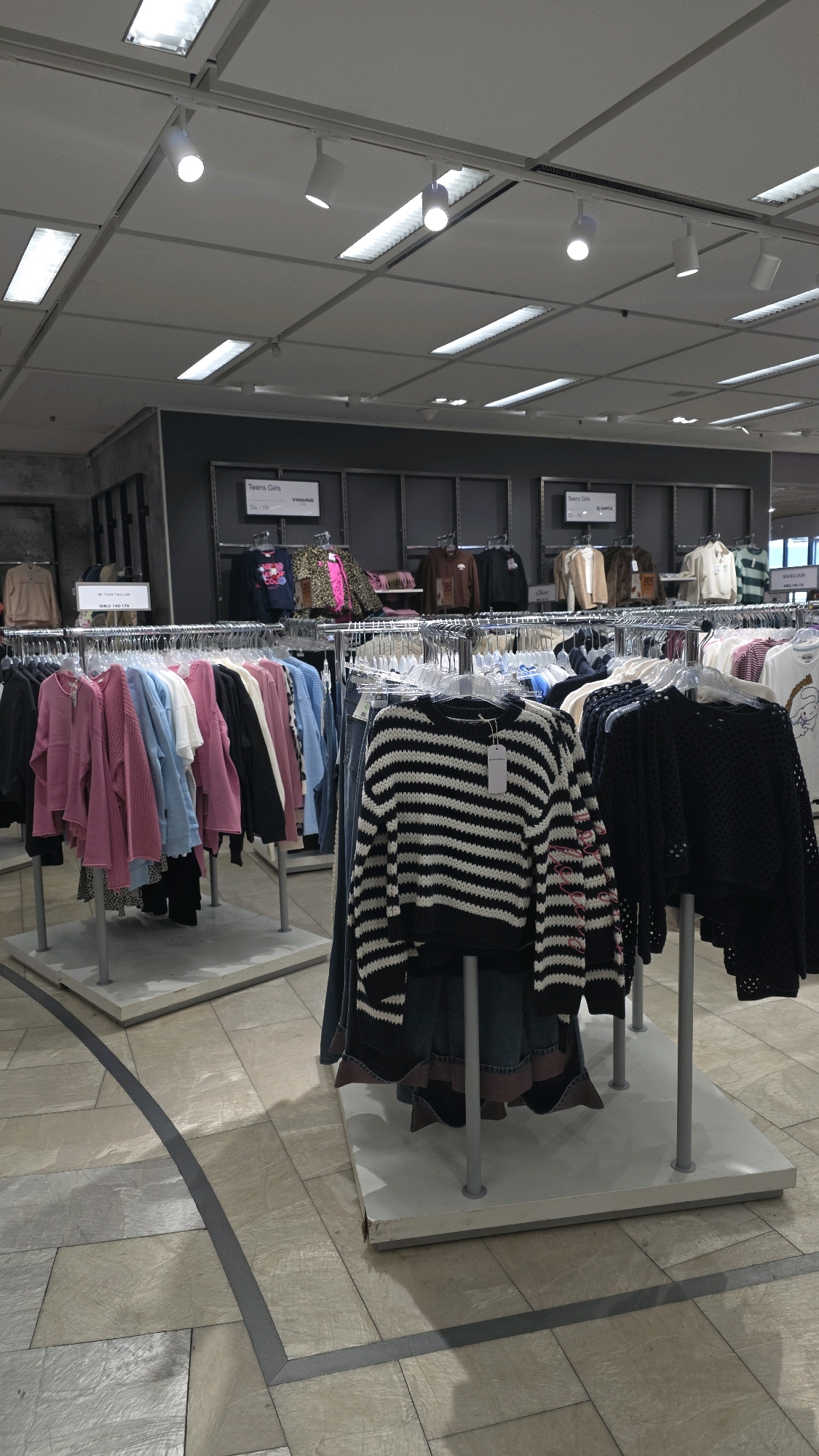 This is a photograph taken inside a clothing store, likely a department store or outlet, in Bonn, Germany. The image captures a section of the women's or girls' clothing department. In the foreground, several clothing racks are visible, displaying a variety of garments. One rack on the right prominently features a black and white horizontally striped sweater. To its left, another rack holds a selection of pink, light blue, and white sweaters and blouses. Further back, more racks of clothing are visible, with some display signs for brands like "Vingino" and "TOM TAILOR". The store features a dropped ceiling with fluorescent lights and track lighting. The floor is tiled in a light beige and grey pattern. There are no people visible in the image.