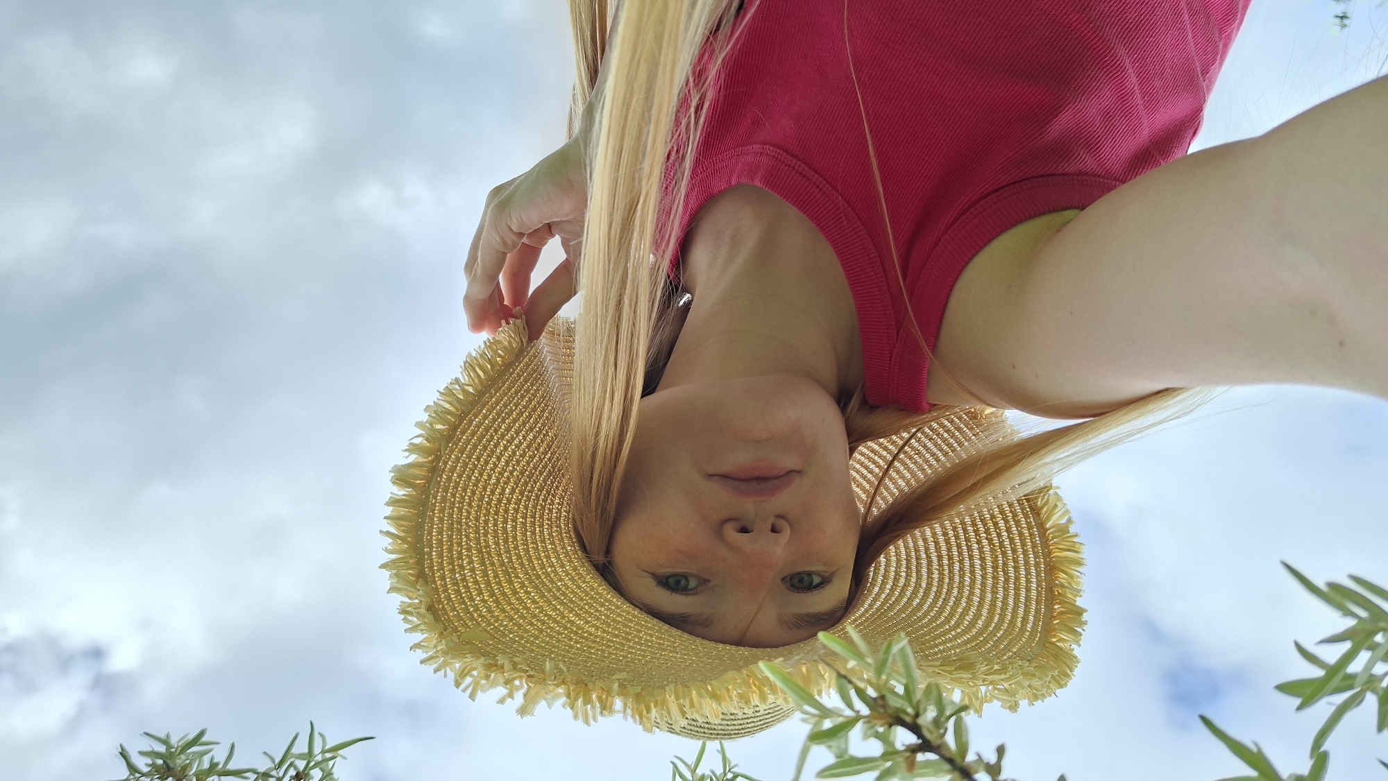 A young woman with blonde hair, wearing a straw hat and a pink tank top, is looking up at the camera. Her head is tilted slightly, and she is holding the brim of her hat with her right hand. The background is a cloudy sky with some green foliage in the foreground, suggesting an outdoor setting. There are no visible indications of the city of Большой Букор, Russia.
