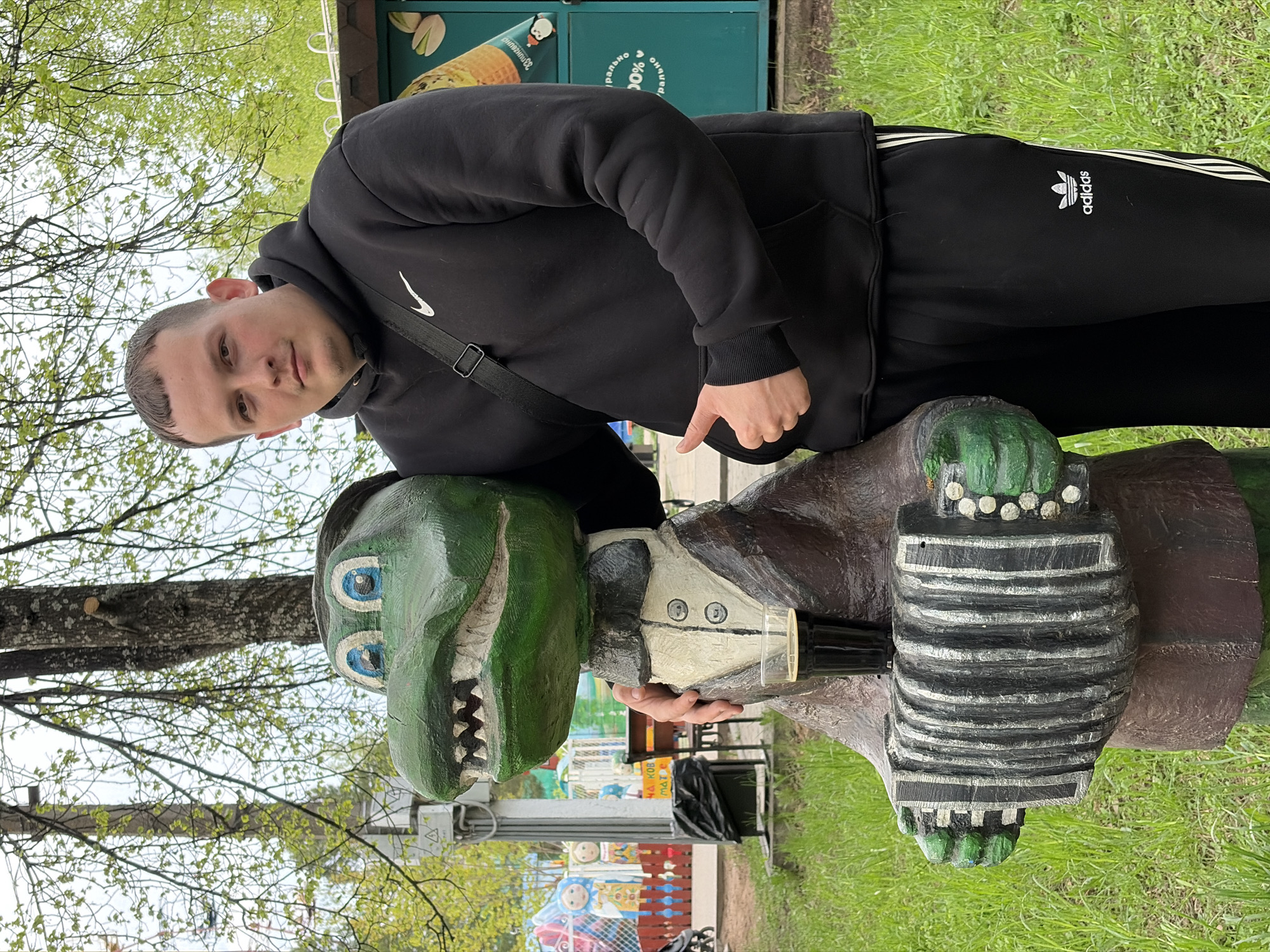 A waist-up shot shows a man resting his left arm on a wooden sculpture of a green crocodile dressed in a brown suit and holding a black accordion in Chaikovsky, Russia. The man, who is white, has short dark hair and is wearing a black hooded sweatshirt with a white Nike swoosh on the left chest, black Adidas sweatpants with three white stripes down the side, and a black shoulder bag. He is leaning his head towards the camera, looking at the viewer. The crocodile sculpture has a wide mouth with small, painted white teeth and blue eyes with white and black detailing. The sculpture appears to be in a park-like setting with green grass, trees, and other structures visible in the background. A green container, possibly for waste, is visible behind the man. The sky is overcast.