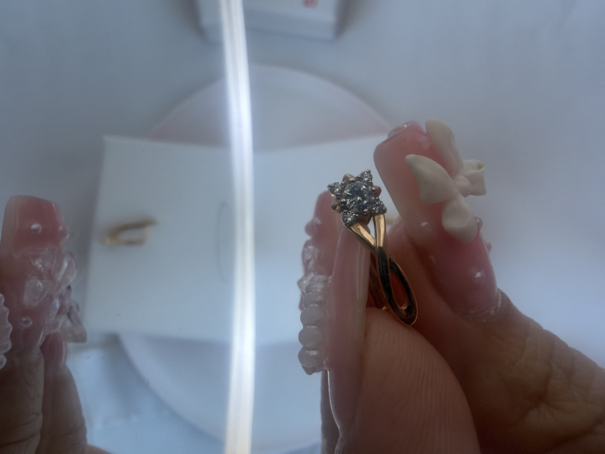 In this close-up, dimly lit, and overexposed image, a gold ring with a diamond cluster is held by a person's hand with long, decorated fingernails. The ring is positioned in the foreground, with its intricate design prominently displayed. The background is out of focus, showing a white surface and a hint of what appears to be a jewelry box. The lighting is harsh, creating bright highlights and washed-out areas, which obscures many details of the environment. The image appears to be taken indoors in an unknown location.