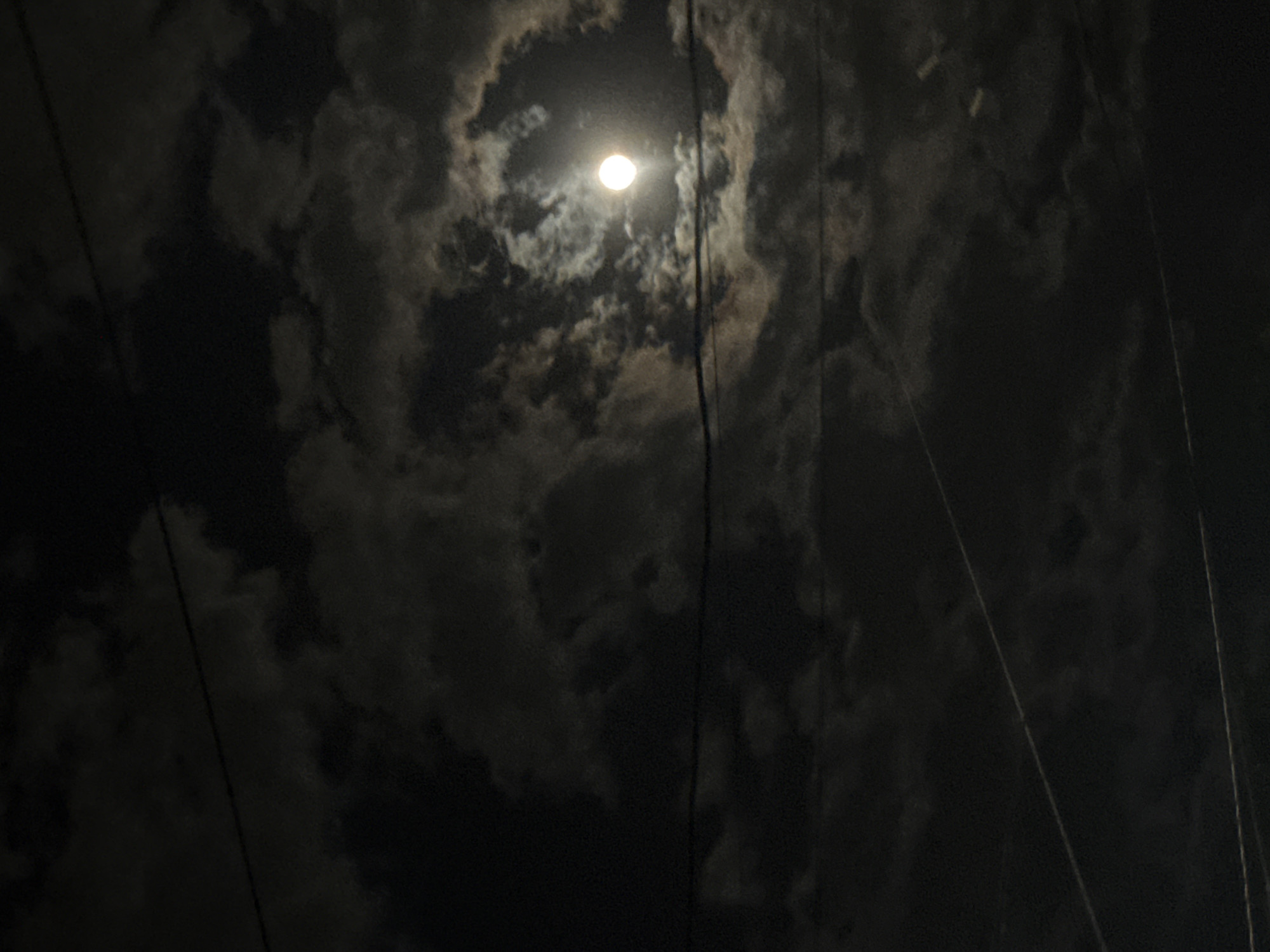 The image shows a night scene in Yerevan, Armenia. A full moon shines through a break in the cloud cover. Power lines are visible in the foreground.