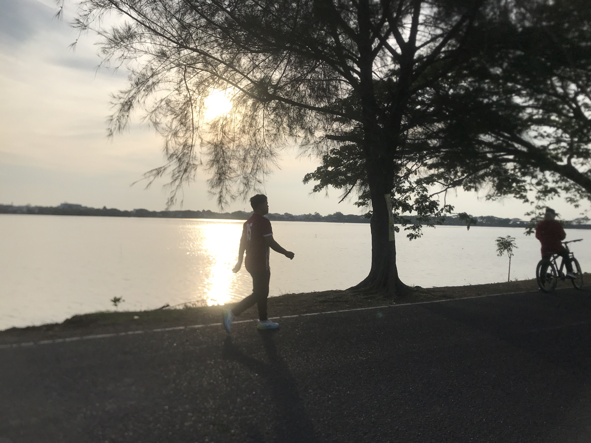 The image captures a scene at Waduk Jeulikat, a reservoir located in Lhokseumawe, Indonesia, during the late afternoon or early evening. The sun, low on the horizon, casts a bright reflection on the water's surface.
In the foreground, a person is walking along a paved path. They are wearing a red short sleeve shirt with a white number "15" on the back, black pants, and white sneakers. A cyclist on a bicycle, wearing a red jacket, is further down the path, heading in the same direction.
A large tree dominates the composition, its branches partially obscuring the sun and casting shadows on the path. The reservoir stretches out into the distance, with the distant shoreline visible under the bright sky.