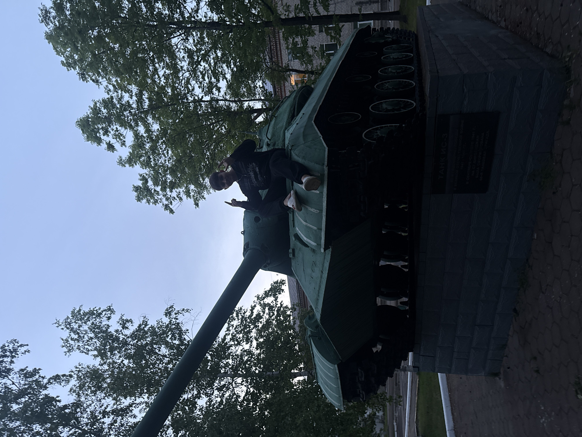 The image displays an outdoor scene in Birobidzhan, Russia, featuring a young person sitting on a historical tank monument. The person, dressed in dark clothing including a hoodie and pants, with light sneakers, is casually posing on the tank's hull, making a gesture with one hand.
The central object is a large, dark green tank, identified by a plaque on its pedestal as a "ТАНК ИС-3" (TANK IS-3). The tank is mounted on a substantial grey, brick-patterned stone base. The plaque, written in Cyrillic, includes text such as "В ВЕЛИКОЙ ОТЕЧЕСТВЕННОЙ ВОЙНЕ" (In the Great Patriotic War), "1941," and "В СЕНТЯБРЕ" (In September), along with phrases like "БЫЛА СФОРМИРОВАНА" (was formed) and "60-Я ТАНКОВ" (60th Tank), likely referring to a tank brigade.
The background features numerous green trees with dense foliage, suggesting a park or memorial area. A multi-story building is partially visible through the trees in the distance. The sky above is a clear, pale blue-grey, indicating either an overcast day or early morning/late afternoon light. The ground around the monument is paved with light and dark grey bricks. The overall composition is taken from a low angle, giving the monument an imposing presence, with the horizon tilted.