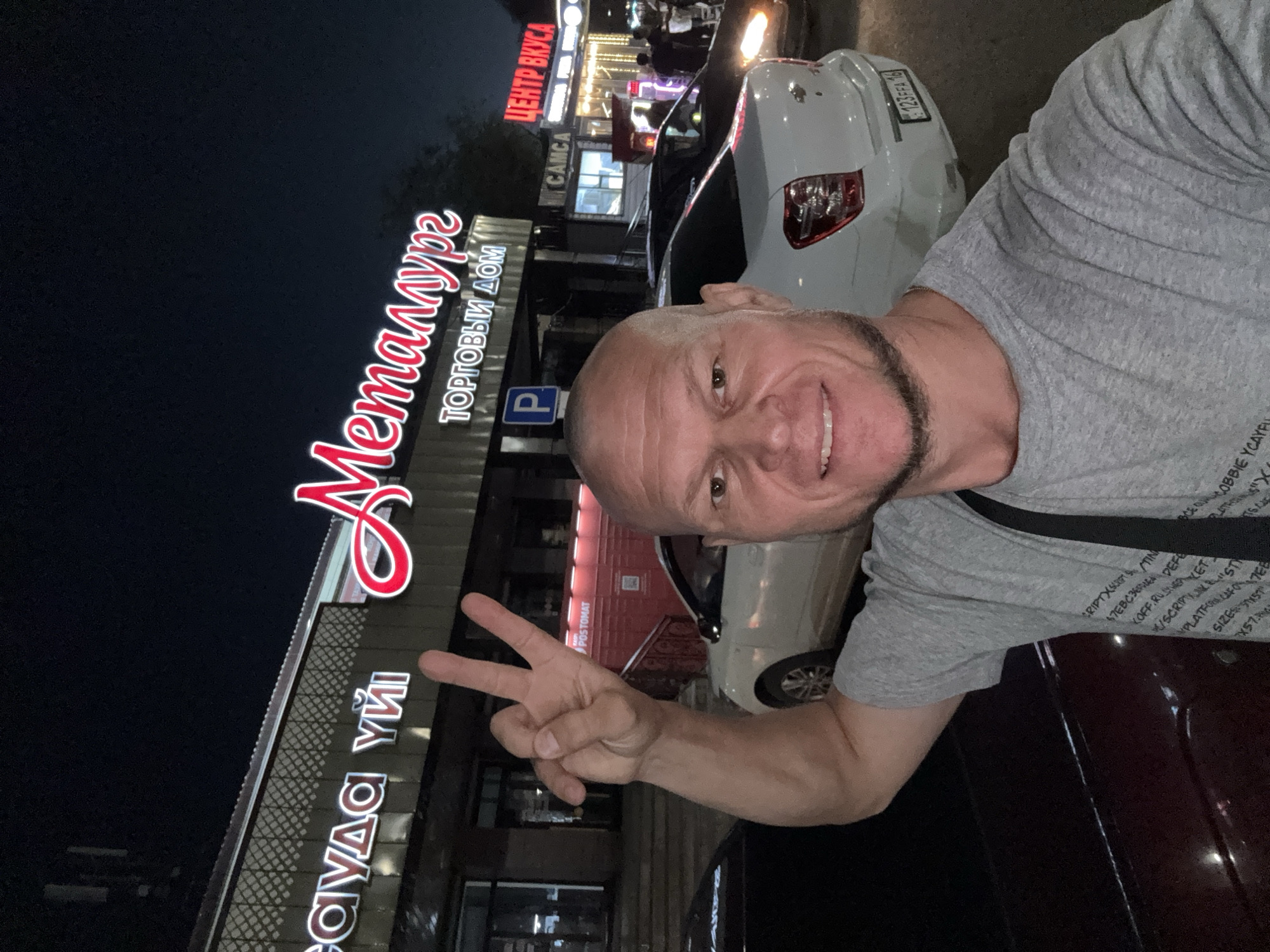 A bald man with a goatee smiles for a selfie at night in front of a building with bright neon signs. The man is wearing a grey t-shirt and has his right arm raised, making a peace sign with his fingers. The building behind him has two signs; one reads "Metalurge" in large red cursive letters with "ТОРГОВЫЙ ДОМ" (Trading House) written below it. Another sign to the right reads "ЦЕНТР ВКУСА" (Center of Taste) in red lettering. A white car with a license plate ending in "16" is parked in front of the building. The environment appears to be urban, with streetlights and other buildings visible in the background. Based on the architecture and signage, the location is likely Oskemen, Kazakhstan.