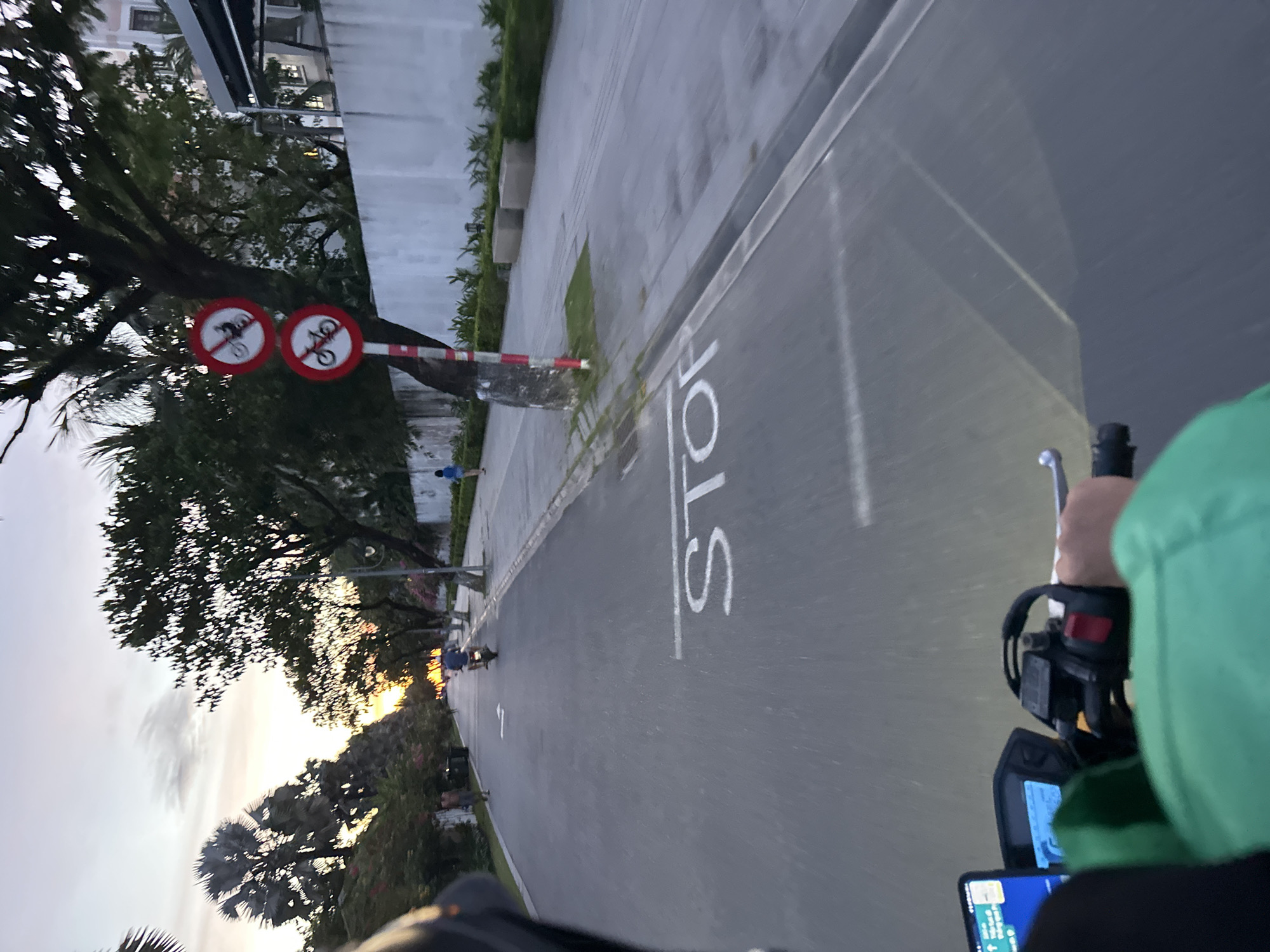The image is a first-person view from a motorcycle in Da Nang, Vietnam, at dusk. The road is paved, with a white "STOP" painted on it. On the right side of the road, there is a sidewalk and a white wall. A few trees are visible, including palm trees. There are two circular signs on a pole, one with a motorcycle symbol crossed out, and another with a bicycle symbol crossed out, indicating no motorcycles and no bicycles are allowed. Several people are visible on the road and sidewalk, including a person riding a motorcycle in the distance, and a child walking on the sidewalk. The sky is a soft orange and gray, suggesting the sun is setting. The rider is wearing a green jacket and has a phone mounted on the handlebars.