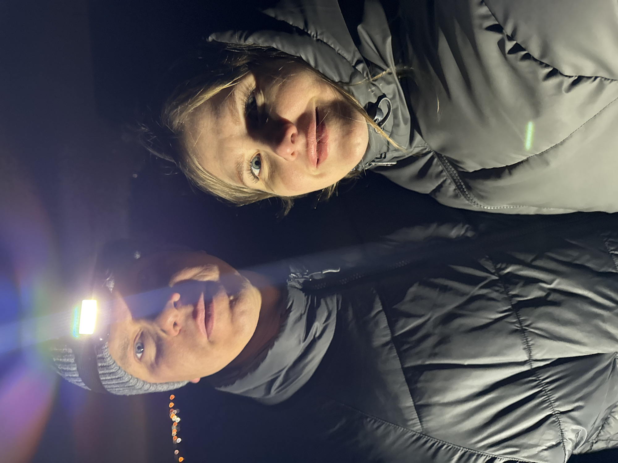A nighttime photograph shows two individuals in an outdoor setting. The person on the left wears a grey beanie and a headlamp, the beam directed forward. The person on the right is positioned above the other. Both are wearing puffy jackets. The background is dark, with some blurred lights visible at the bottom left. The location is not apparent from the photo itself. There is no way to ascertain the location is Горный, Russia.