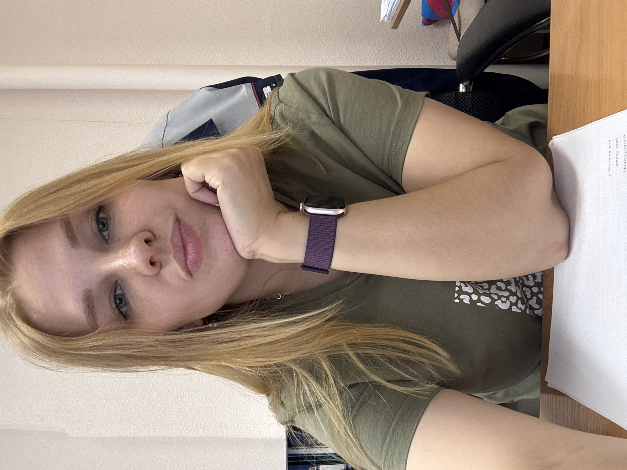 The image shows a young Caucasian woman with blonde hair, wearing a green t-shirt and a purple smartwatch, sitting at a desk in an office. She is resting her chin on her hand and looking towards the camera with her lips slightly pursed. The background suggests an indoor environment with a wall, a chair, and some shelves with books. The desk has a sheet of paper on it. The lighting indicates an indoor setting, likely during the daytime. The overall impression is a casual, possibly work-related photo. The location of Заволжье, Russia, is not visually verifiable from the image itself.