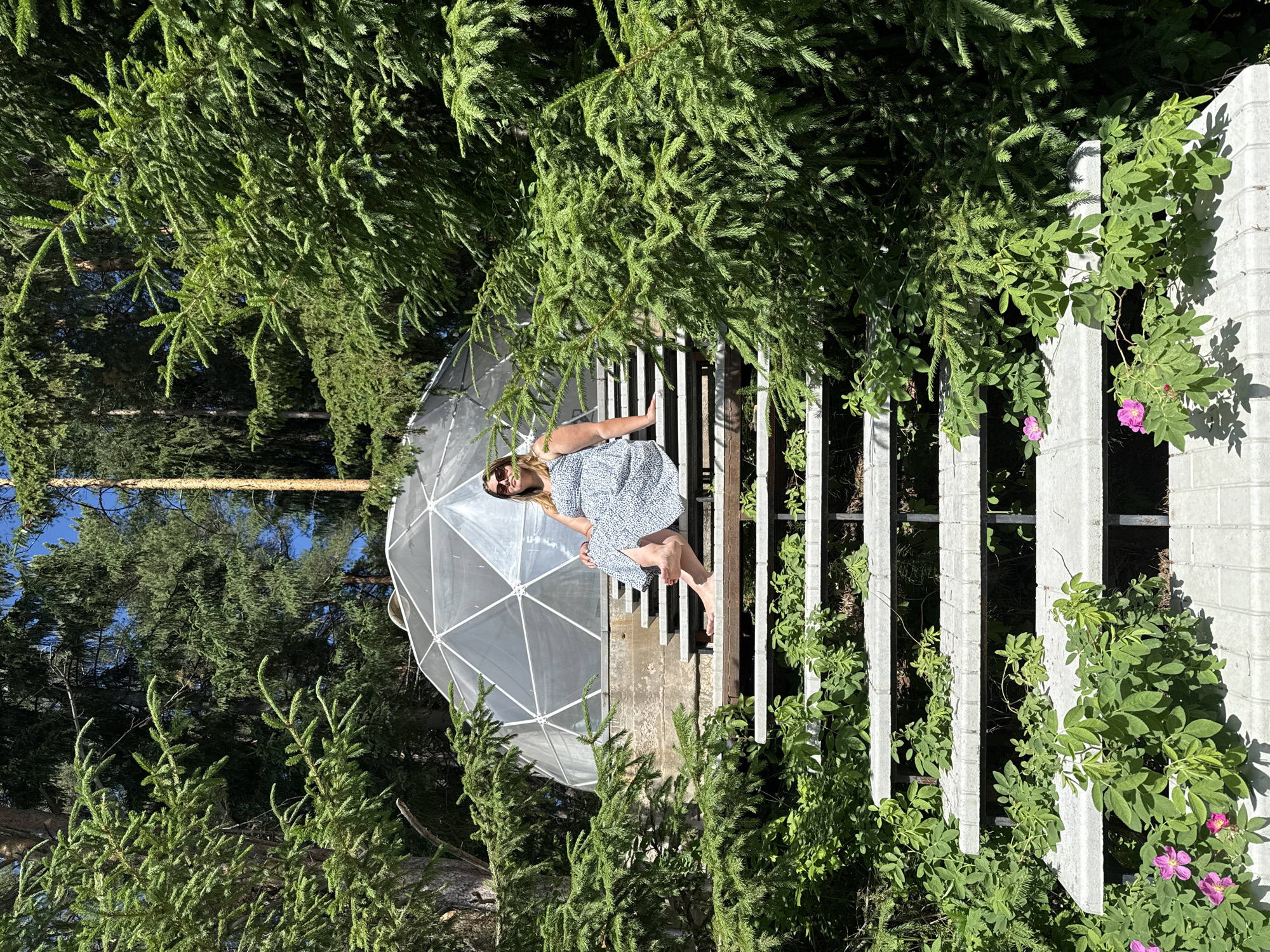 In Makarovo, Russia, a woman sits on the steps of a concrete staircase outdoors. She is wearing sunglasses and a blue and white patterned dress. The staircase is surrounded by lush green trees and vegetation, and behind it, a geodesic dome structure is visible. The sunlight suggests it is daytime.