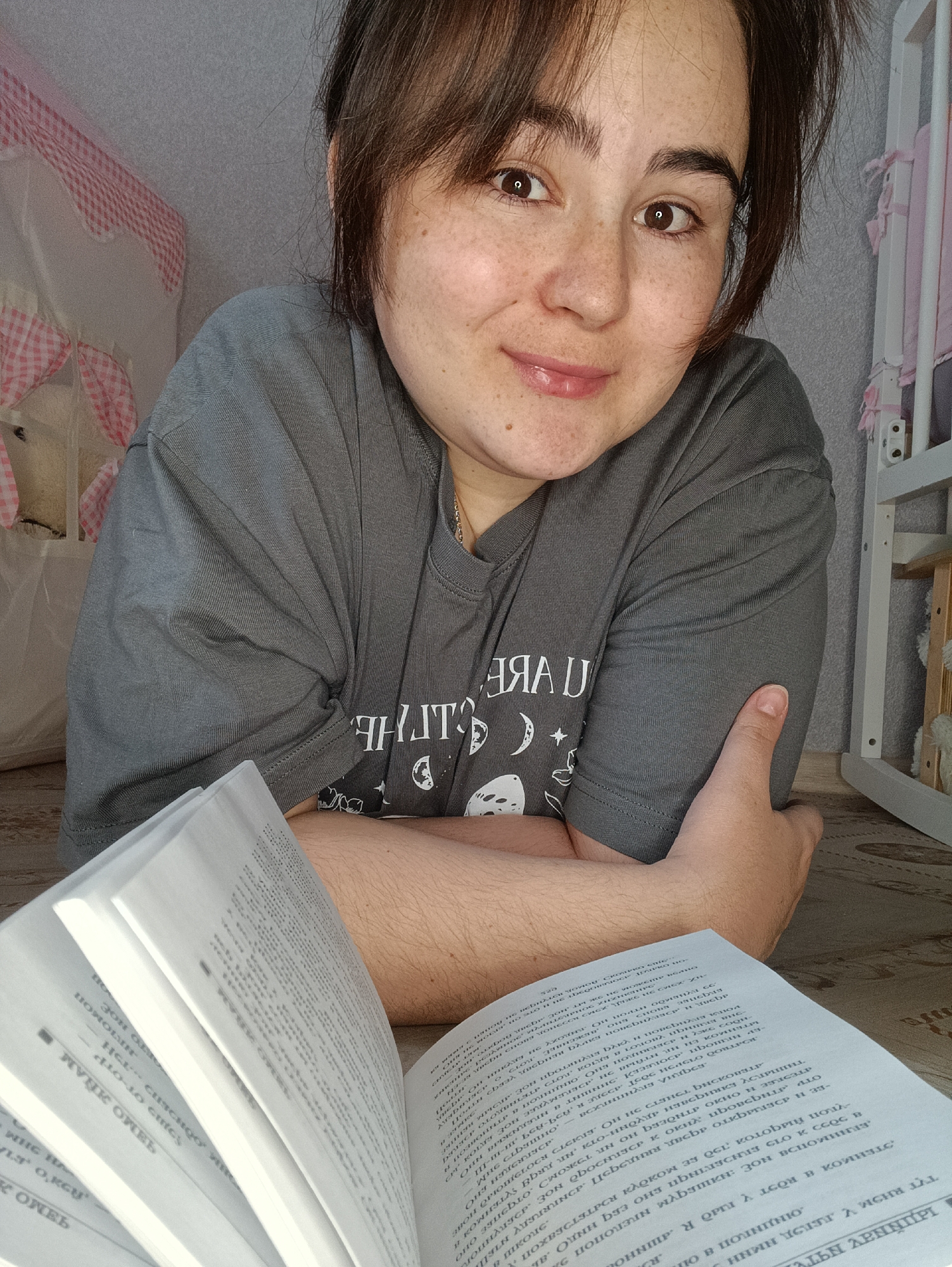 The image shows a woman with brown hair and freckles lying on the floor, propped up on her arms. She is wearing a gray t-shirt with white writing and designs. In front of her is an open book with Russian text. In the background, there is a child's crib with pink and white gingham fabric. The wall is a light neutral color. The floor appears to be a patterned surface. The location cannot be verified from the image, and the OCR text does not provide additional information.