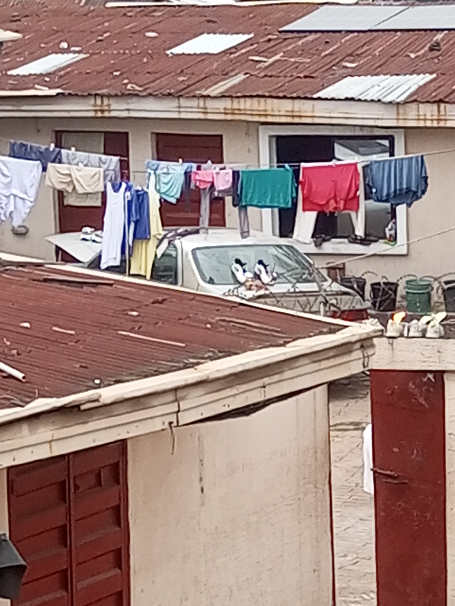 The image presents a residential scene in Onitsha, Nigeria. A multi-story building features a rusty corrugated iron roof with interspersed white patches. Laundry, including clothing items in various colors, hangs on a clothesline strung across the front of the building, obscuring a parked silver car. On the car's hood are two pairs of shoes. Additional pairs of shoes rest on the ledge of a wall to the right of the car, near several buckets. In the foreground, a smaller building with a red corrugated iron roof is visible, its wall painted with red trim. A partial view of a person wearing white can be seen walking outside to the side of this building.